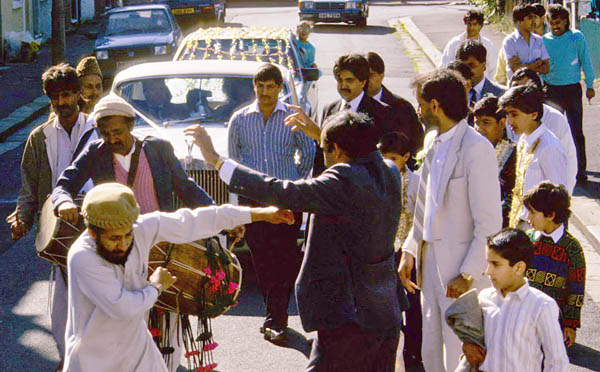 Colour photograph showing a crowd of men and boys on the street.  One is playing a drum and two are dancing.