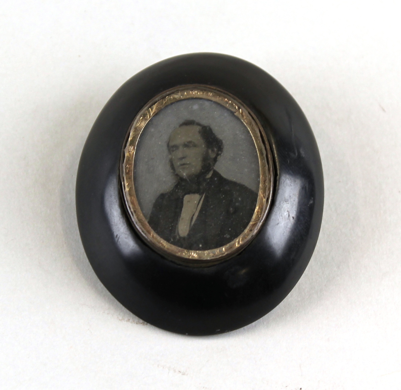 An oval shape brooch with an image of a man, with a gold rim and a black surround. The man is wearing a dark suit and bow-tie.