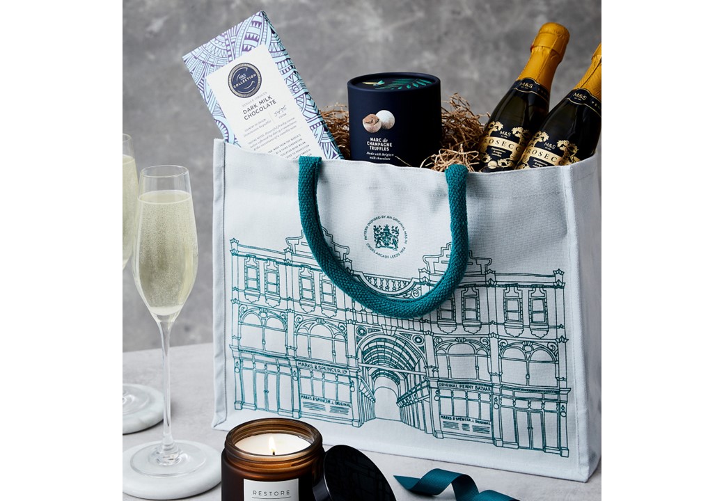 A M&S gift bag with an architectural print contains champagne, chocolate, and truffles surrounded by straw. Two champagne glasses and a lit candle add a celebratory feel.