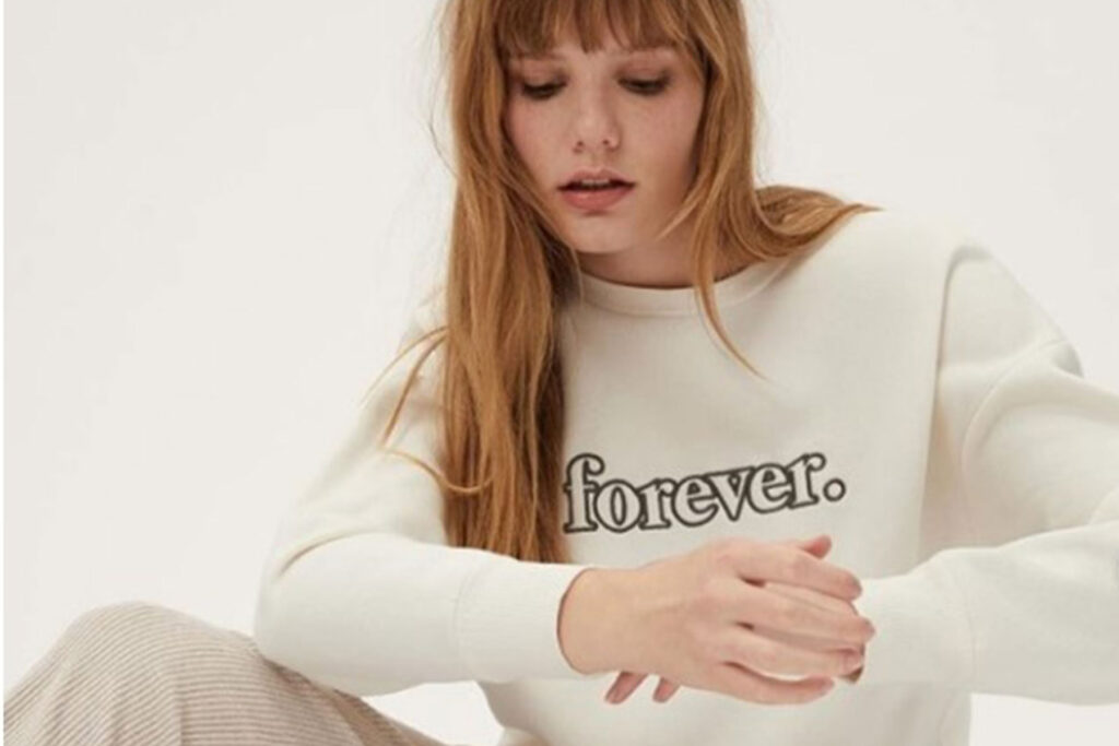 A woman wearing a cream sweatshirt with the word “forever.” sits against a plain light background.