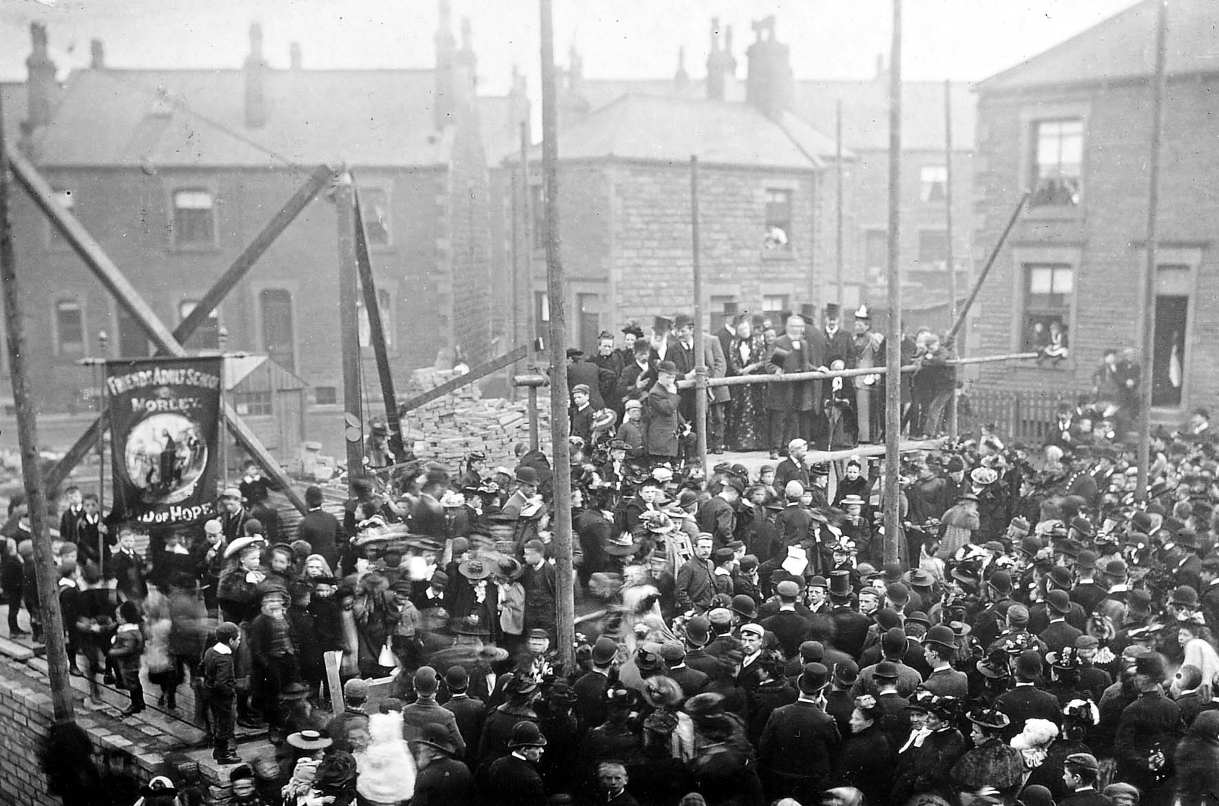 Black and white photo showing a large crowd. There is a smaller crowd of people up on a wooden platform.