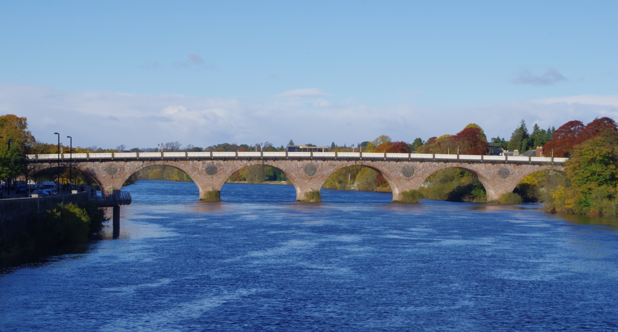 A photograph with bright blue water and a stone bridge half way in the image. A bus is traveling along the bridge. Trees in autumn can be seen on both sides of the bridge. Light blue skies above.