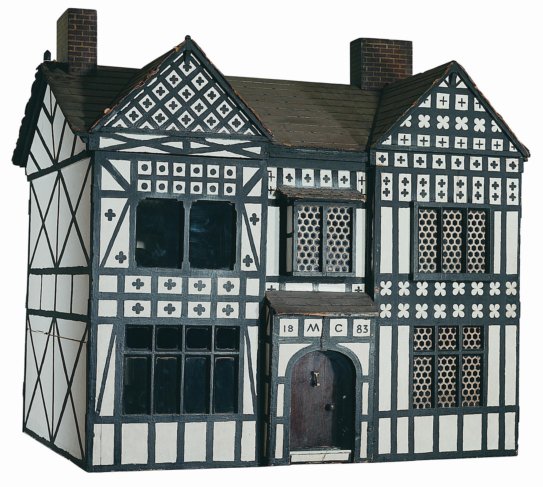 A large, two-storey Doll's House in a black and white Tudor style design with timber framing. It has a front door in the centre, with a window either side. On the first floor it has a gable at both sides, a chimney at each end of the roof, and three large windows on the front wall. The date is written as 18 MC 83 above the door.