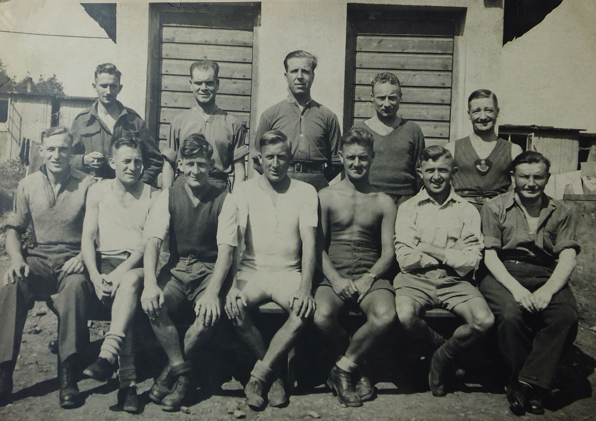 Group photo of thirteen men, likely soldiers, in casual clothing, sitting and standing outside wooden buildings.