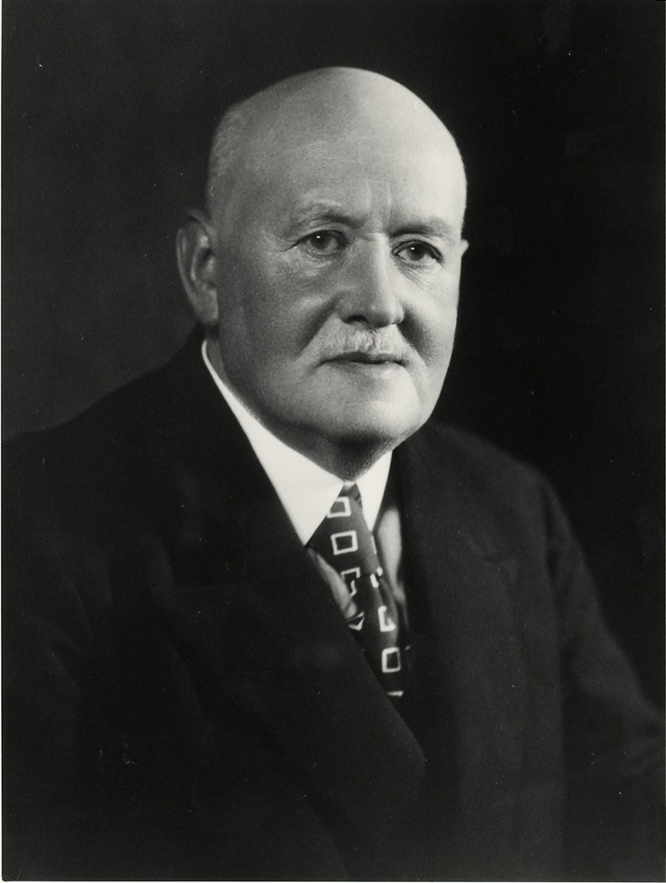 Black and white portrait photo of an elderly bald man wearing a suit and tie