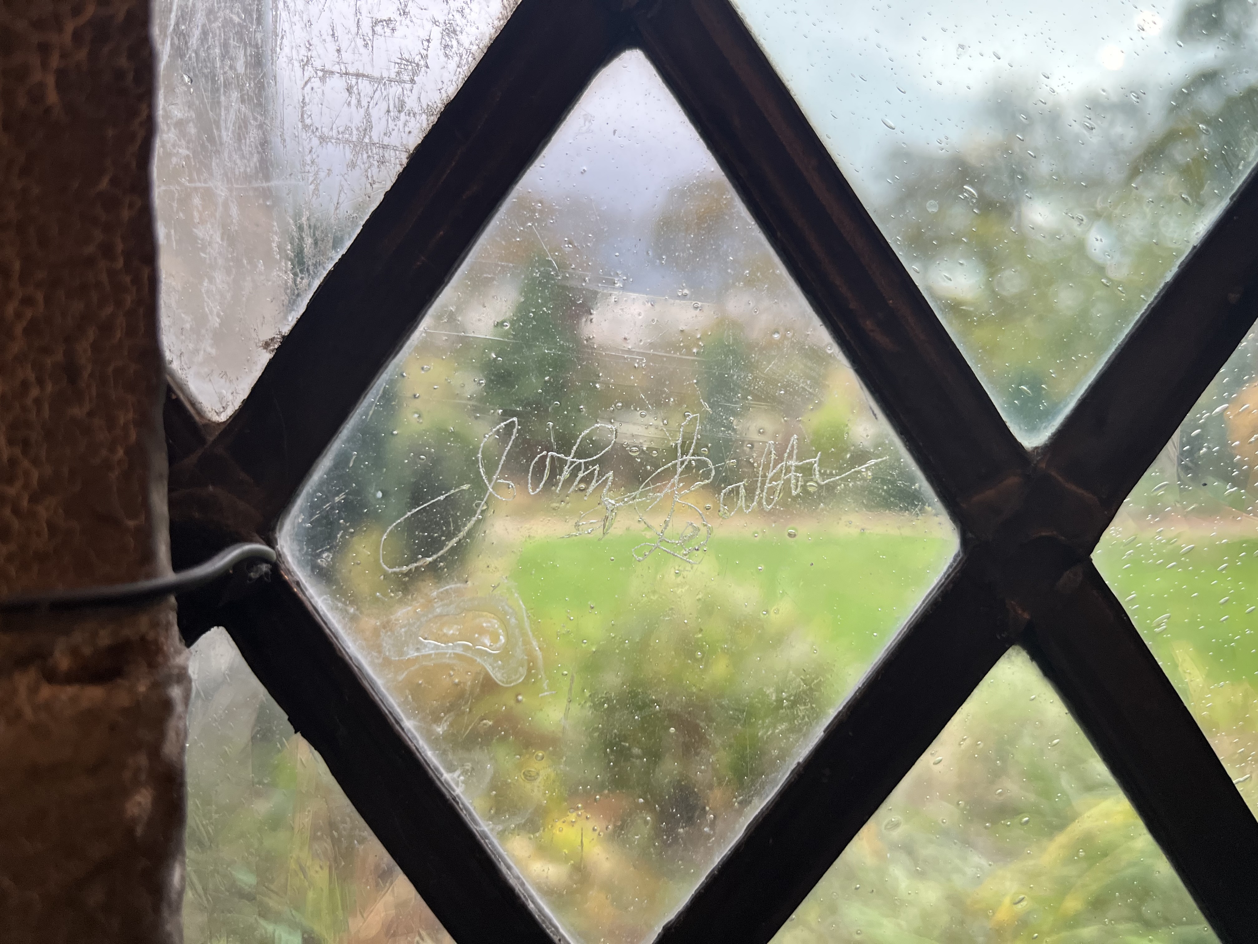 A diamond-shaped windowpane with a signature etched onto it, overlooking a blurred green landscape and trees through the glass.