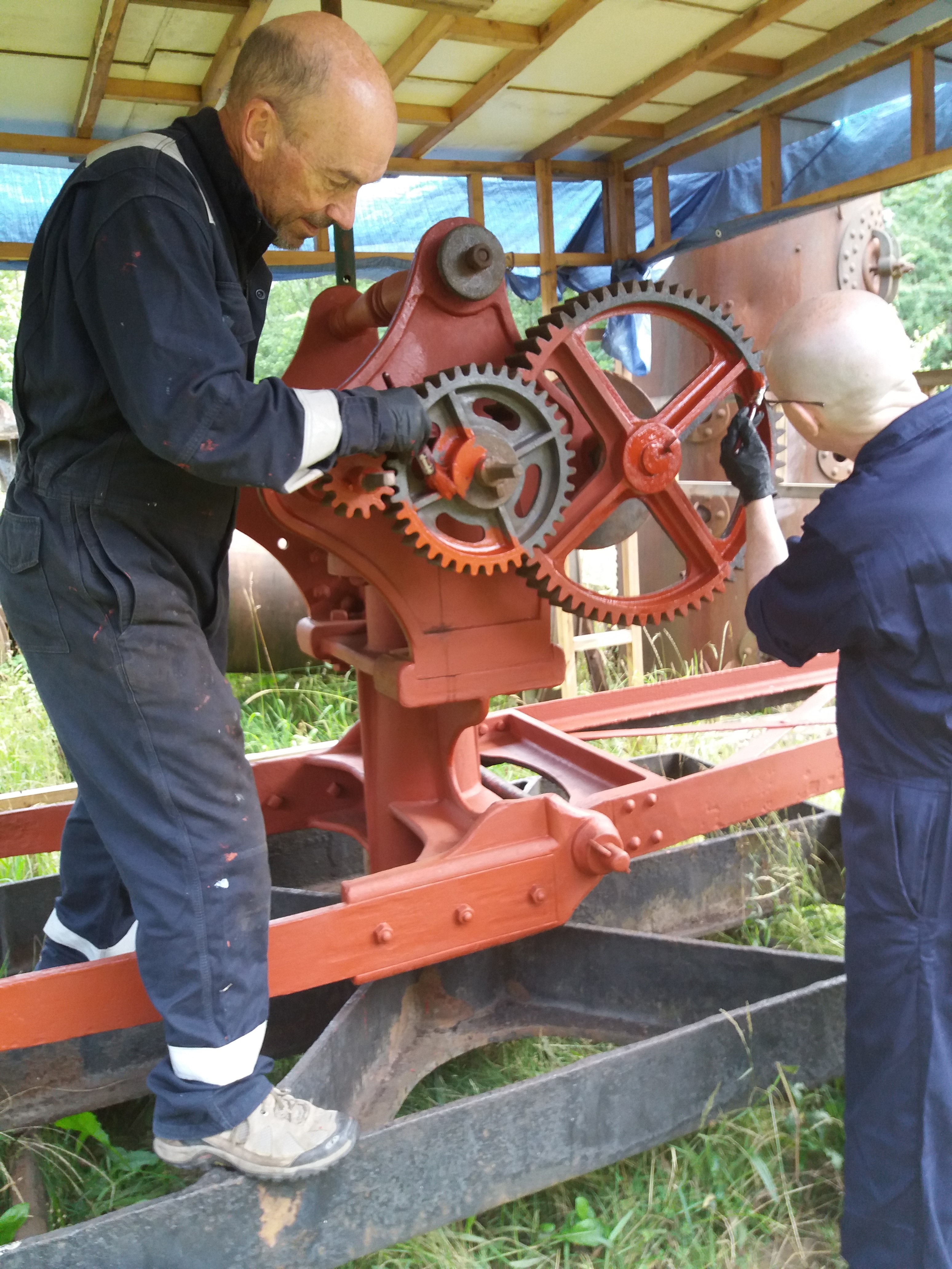 Two men in overalls painting cogs on part of a machine.