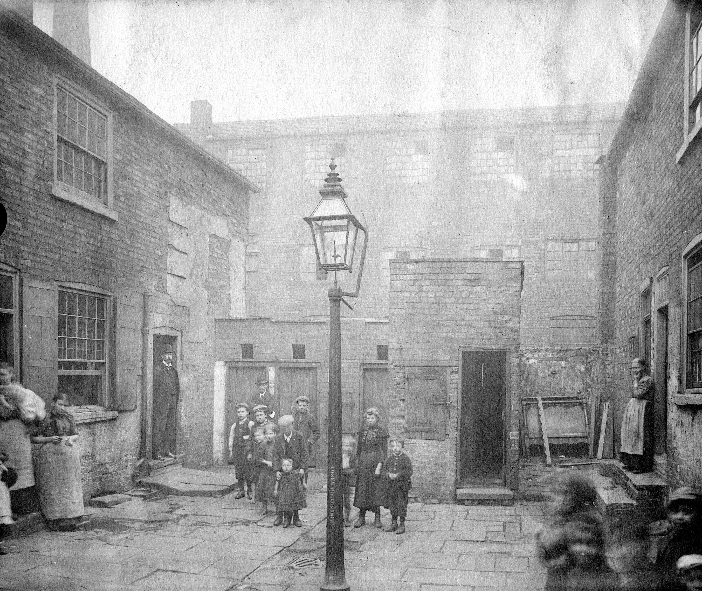 Women and children standing in the middle of a yard with houses on three sides.  Two bowler hatted men can be seen and in the background are the shared toilets.