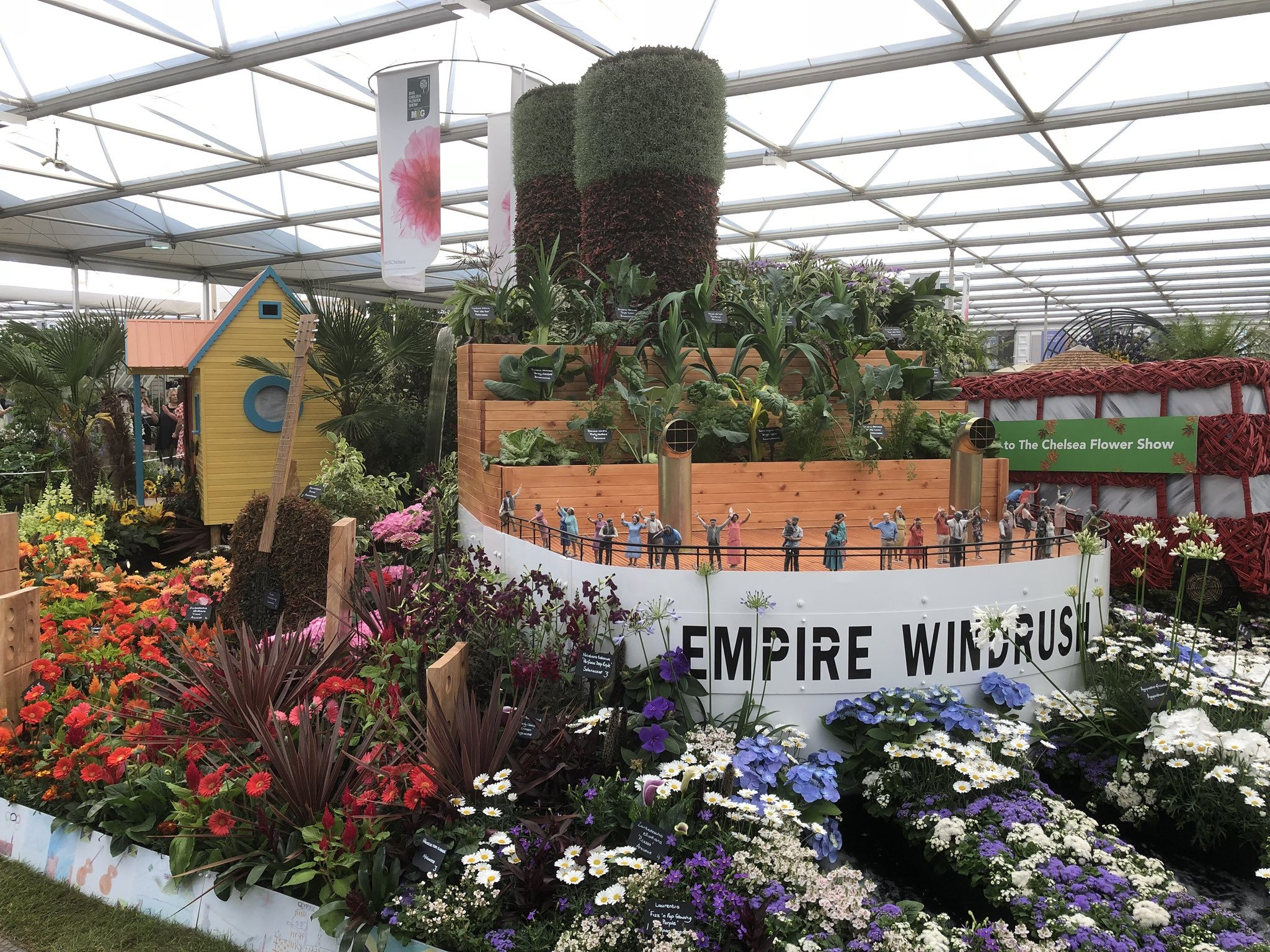 Colour photograph showing a representation of a ship, with chimneys.  On deck are models of people and there are blue, purple and white flowers at the front.