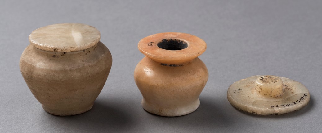 The image shows three small stone jars with flat, round lids. The jars are cream and beige, with one having a reddish tint at the neck. They vary in size, with the leftmost being the largest. The lids are either on top of or next to the jars, all showing smooth surfaces and rounded shapes.