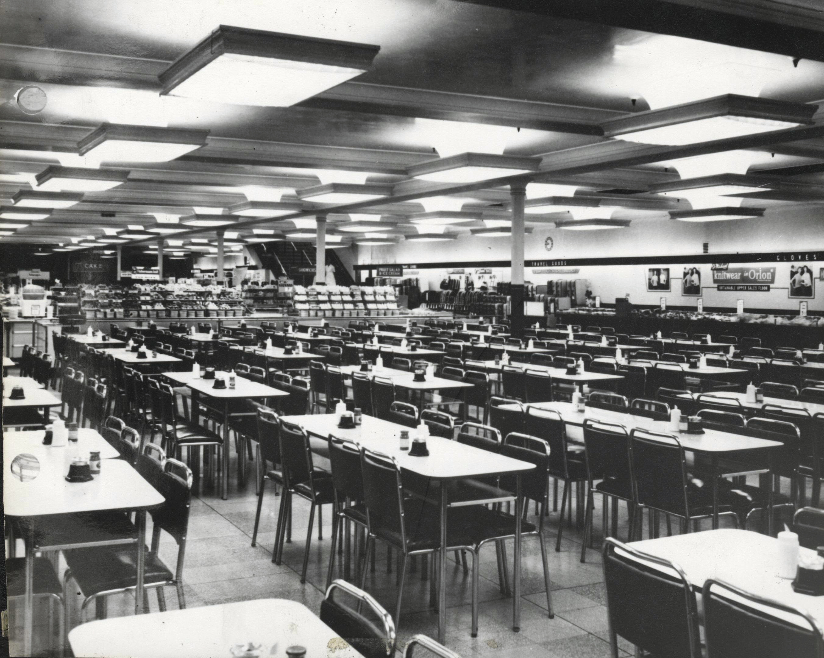 A wide-angle black and white photograph from around 1960 captures an empty Marks & Spencer café bar. The space is filled with neatly aligned rows of square tables and matching chairs, each table topped with condiment sets and napkin holders. Fluorescent lighting fixtures line the ceiling, and in the distance, retail counters and wall signage advertise knitwear and other clothing goods.