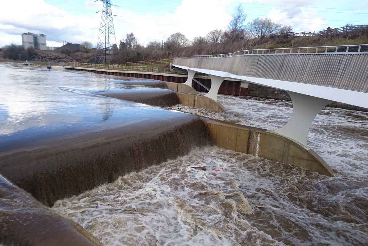 A photograph of a rushing water down a weir and a metal bridge on the far right.