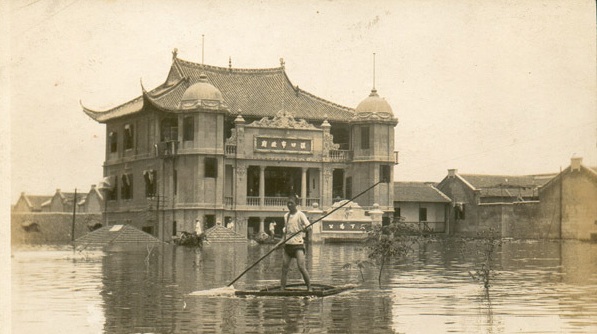Black and white photo of a man standing on top of a piece of wood and punting his way across the flood water.  Behind him is a building, partially submerged underwater.