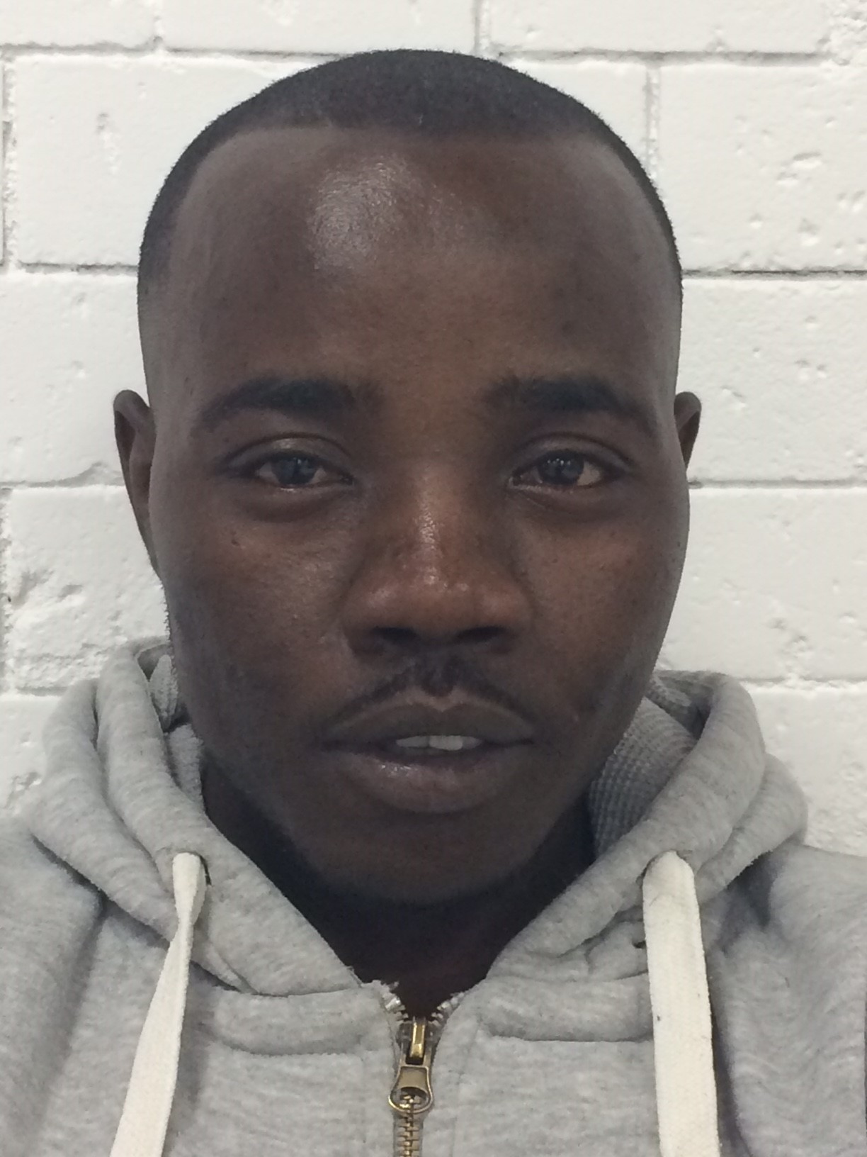 The image is a close-up color photograph of a man standing against a white brick wall. He has a closely shaved haircut and a short mustache. He is wearing a light gray hoodie with a white drawstring and a zipper at the front. The man is looking directly at the camera with a neutral expression. The background is simple and plain, which keeps the focus on his face.