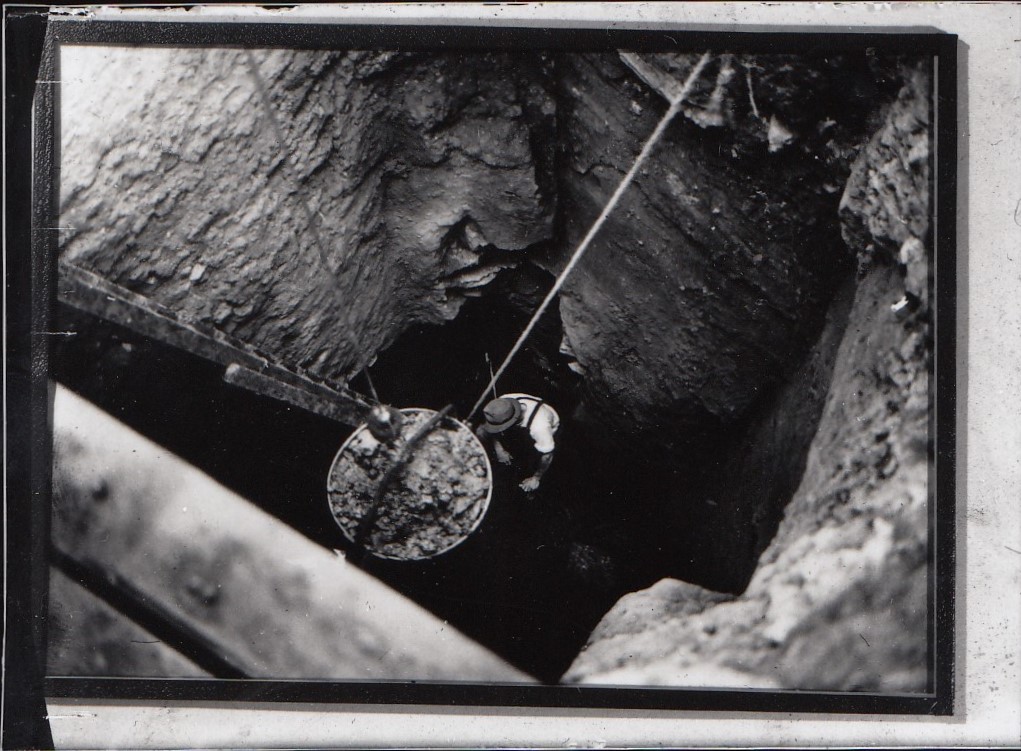 A miner wearing a hard hat is suspended by a rope in a deep, rocky mine shaft. Nearby, a bucket filled with excavated material hangs from a pulley.