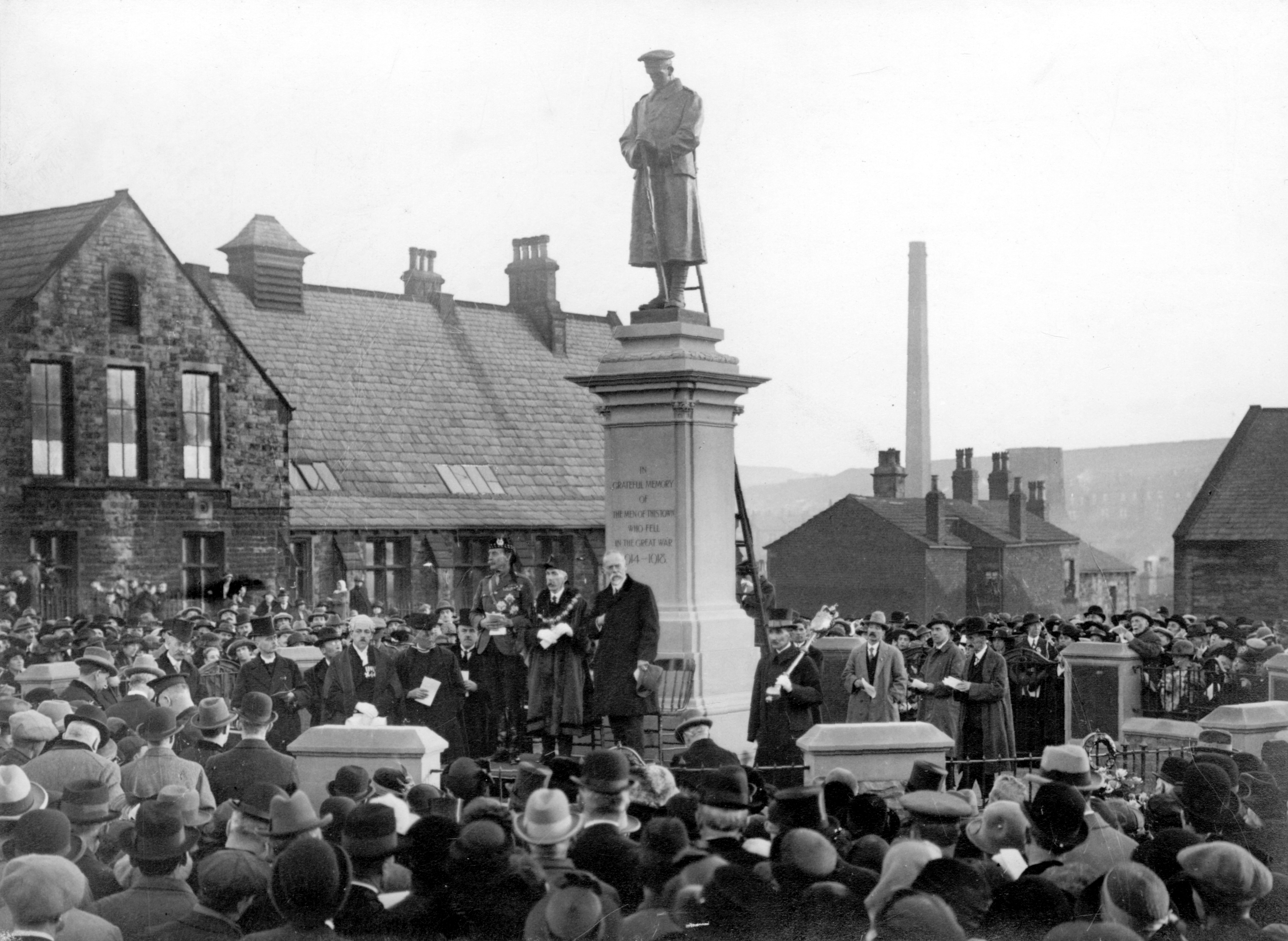 A large crowd gathers around a WWI soldier monument in a town square. Officials stand on a platform, with historic buildings in the background.