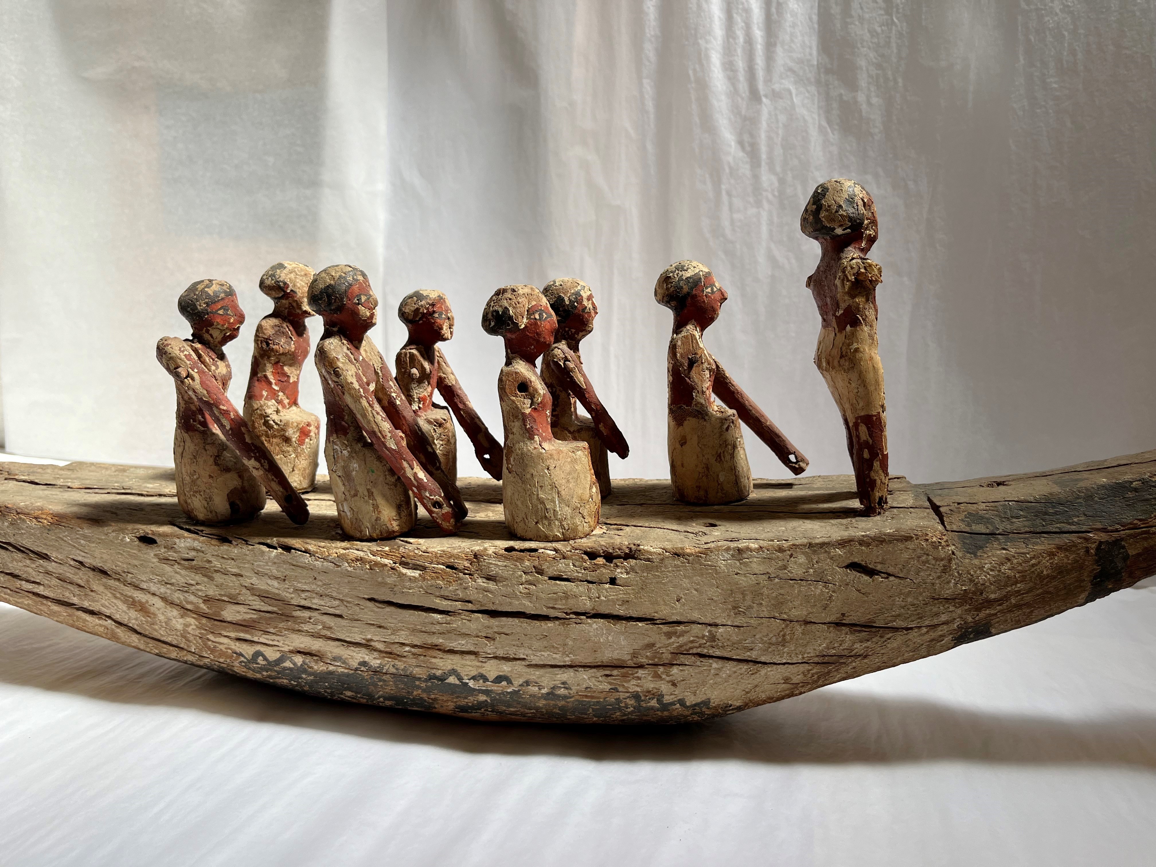A wooden model of an ancient Egyptian boat with eight figures. Seven are seated, rowing, while one stands at the front, possibly a leader. The red and white paint on the figures is worn, and the boat has a curved, weathered hull.