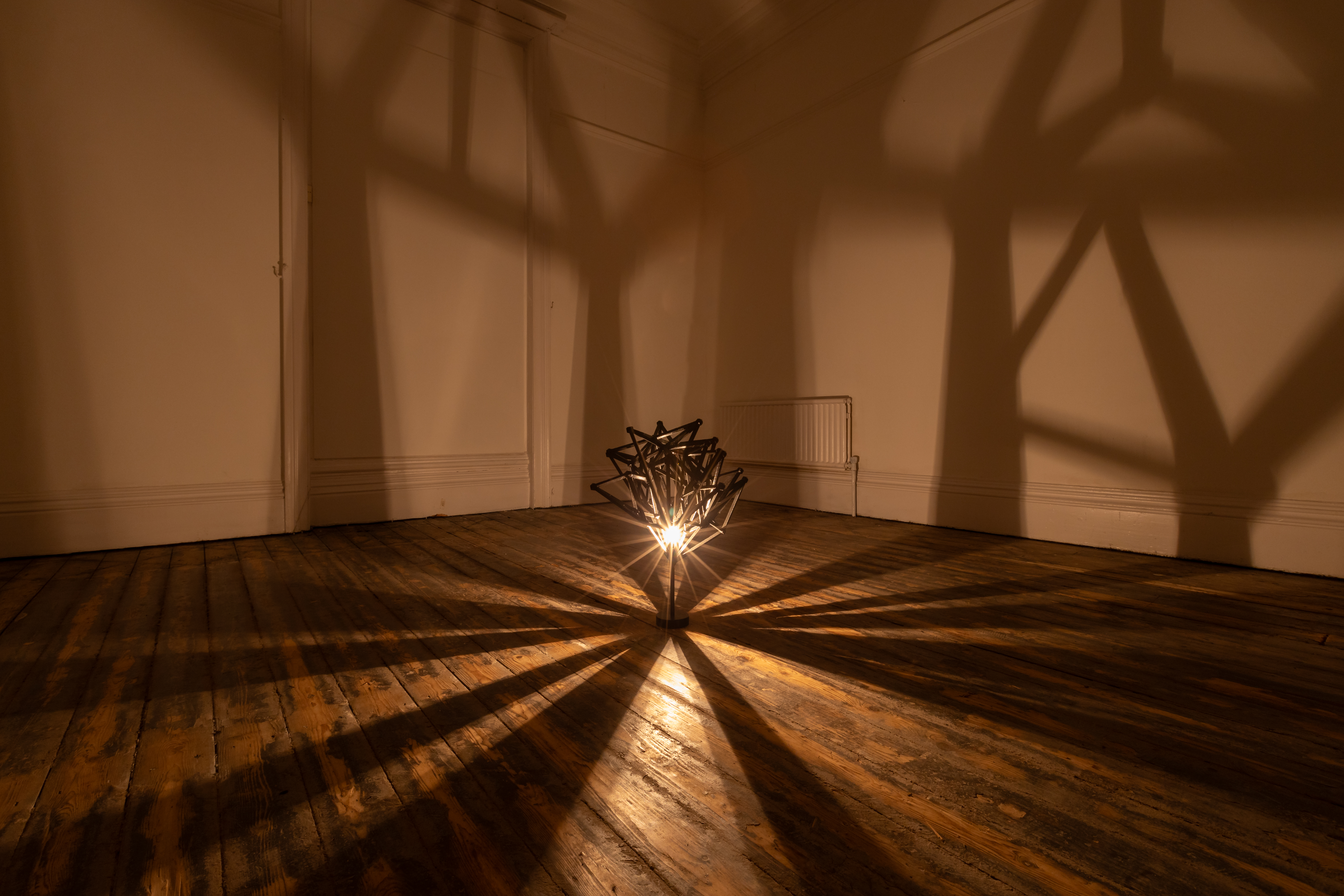 A metal sculpture sits on a wooden floor with light emanating from within.The sculpture is made of many pieces of metallic pieces on a small stand at floor height. Casting shadows onto white walls.
