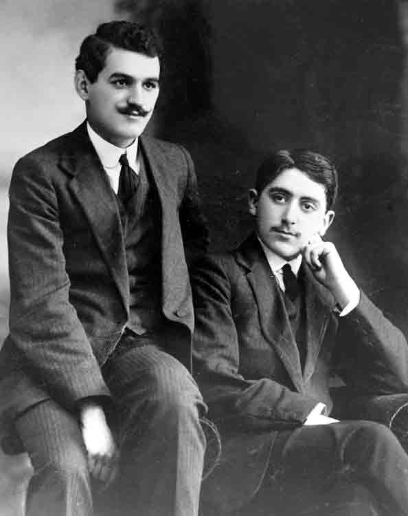 black and white portrait photo of two young men dressed in suits and ties, one seated on chair of arm the other is seated in the chair