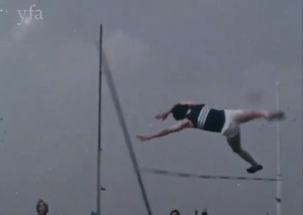 Film still showing a male athlete in mid air going over the pole jump