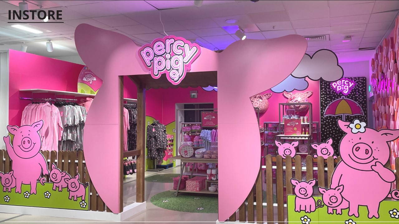 The image shows a vibrant pink Percy Pig-themed store display with oversized pig ears framing the entrance. The setting is playful and inviting, featuring cartoon pigs and colourful merchandise inside.