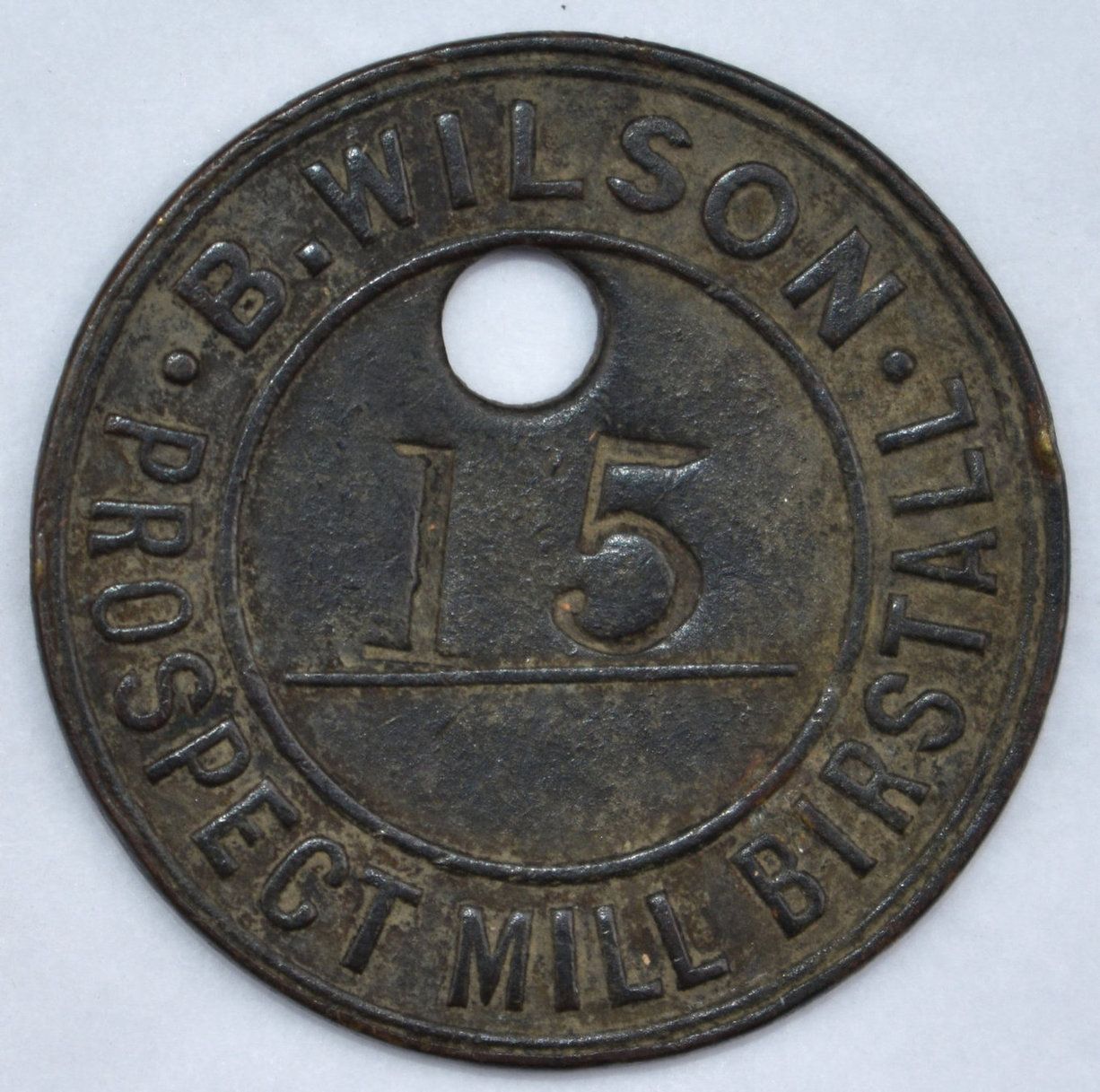 A round, metal mill token with a central hole. The outer text reads "B. WILSON. PROSPECT MILL BIRSTALL" and the centre displays the number "15" above a horizontal line. The token has a worn, aged appearance.
