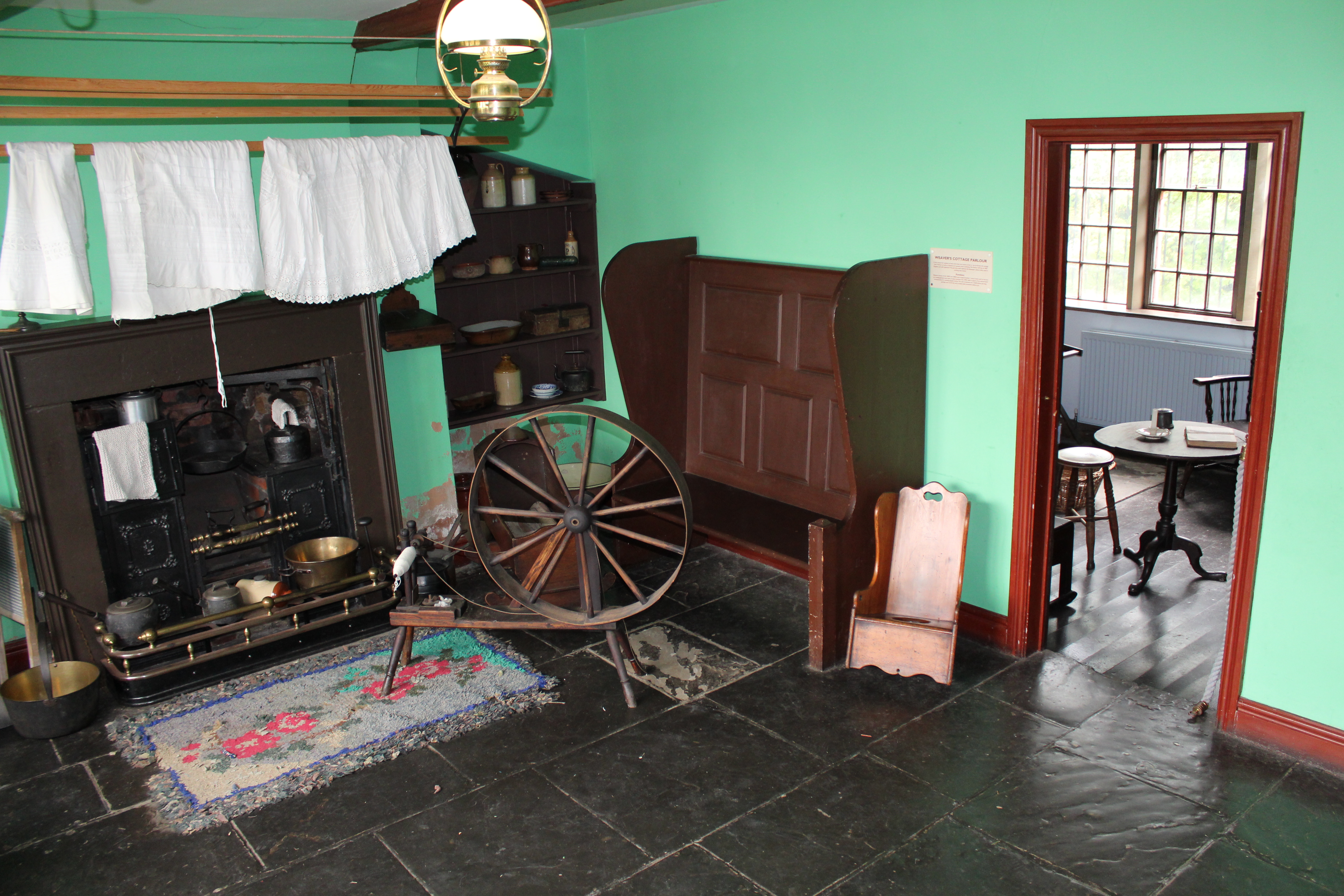 Room with painted green walls, a stone flag floor and a coal fired hearth with a rocking chair in front.  A spinning wheel stands to one side.