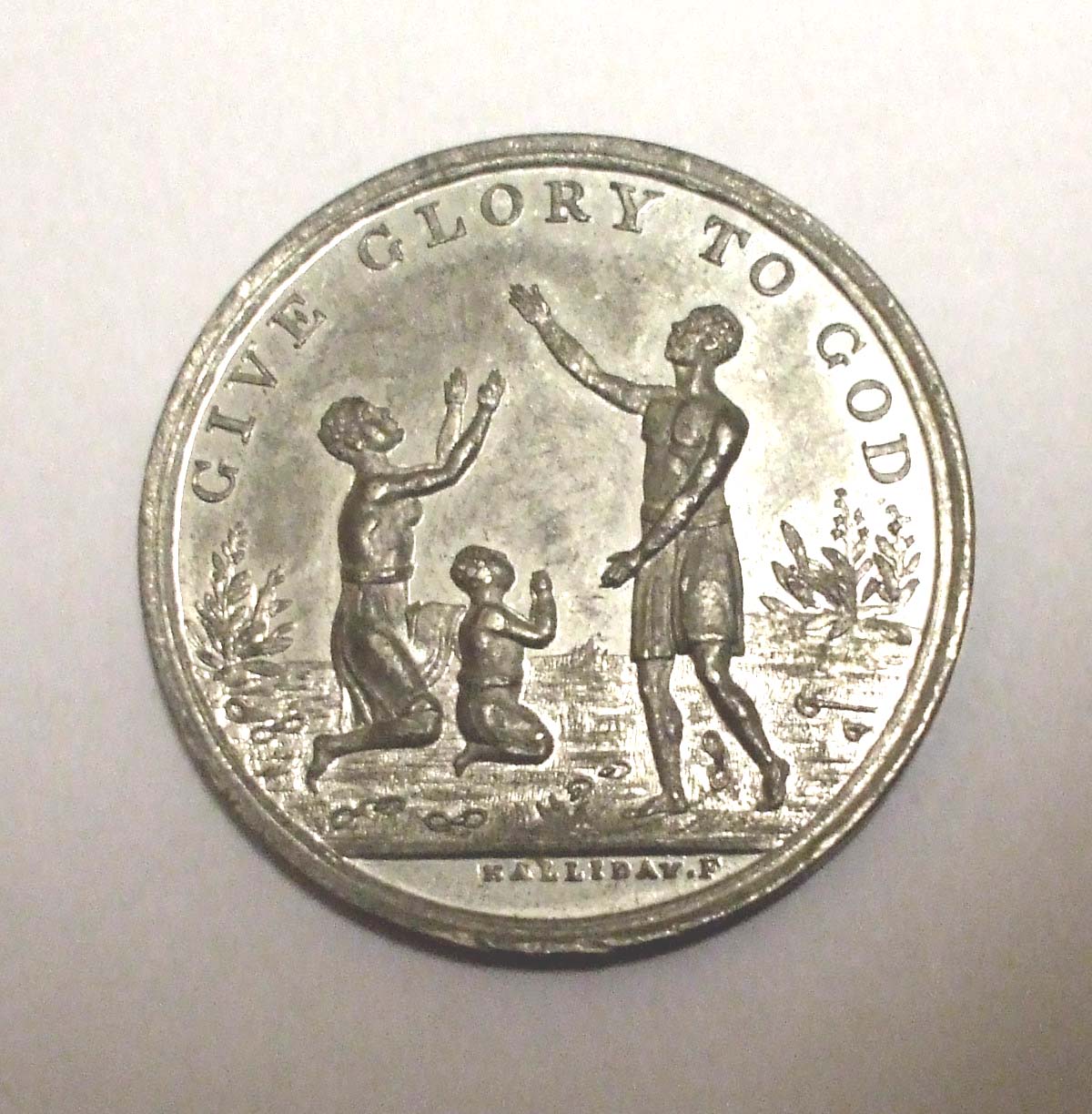 Circular metal medal to commemorate 50 years since the abolition of slavery, with image of a standing man and a kneeling woman and child, arms raised in praise.