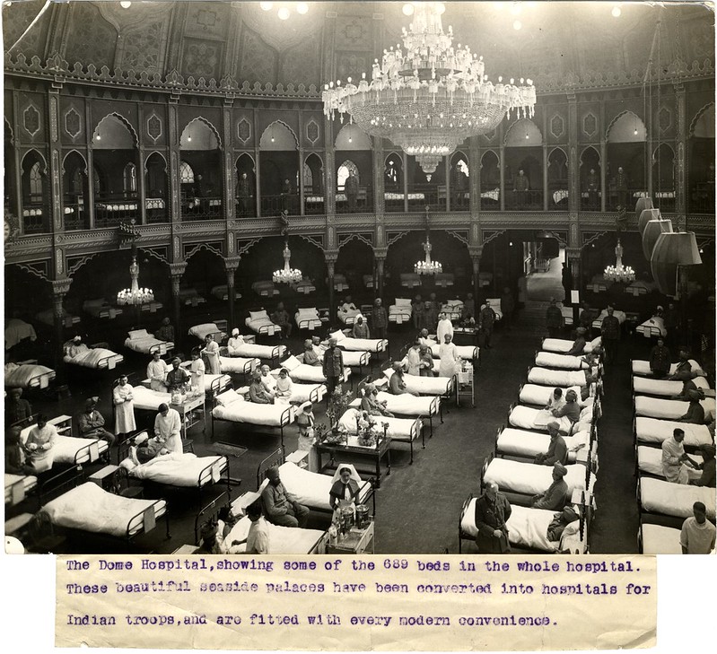 Black and white photograph showing rows of hospital beds and patients. There is a huge chandelier hanging from the ceiling and the building is a theatre.