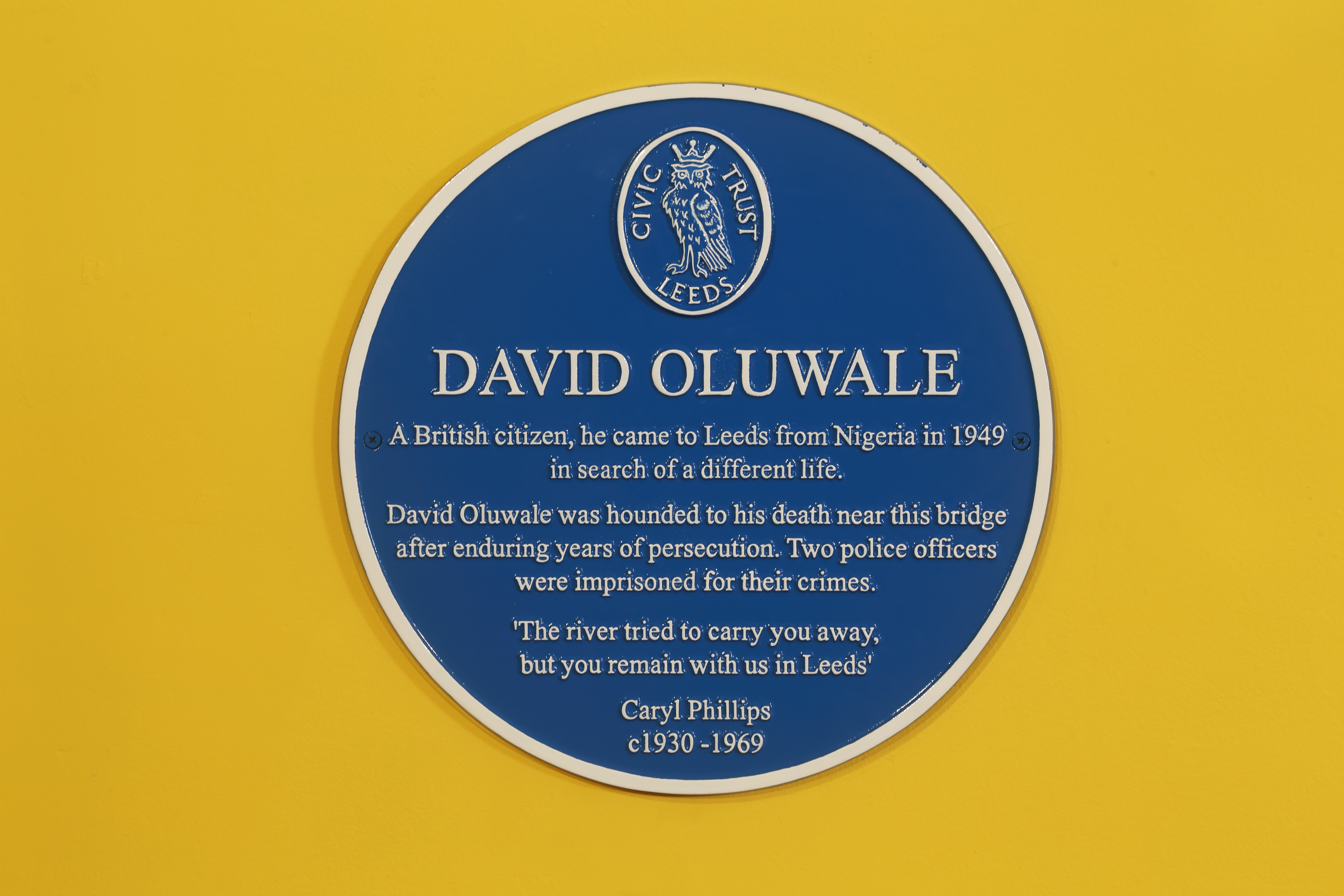 A Blue Plaque for David Oluwale, with the crest for the Civic Trust Leeds and their owl logo. The plaque details that David Oluwale moved from Nigeria to Leeds in 1949, but faced years of persecution and eventually died in the river. His cause of death was unknown, but two police offers were imprisoned for related crimes.