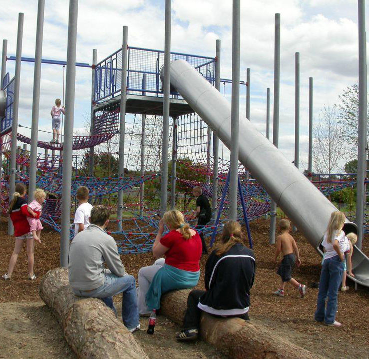 Contemporary photograph of a children's adventure playground, including a bridge, climbing ropes and a tunnel slide.