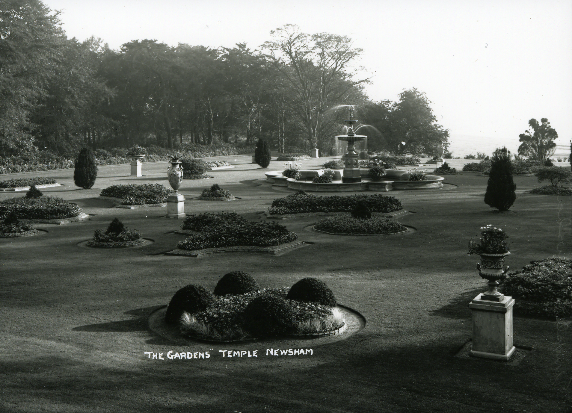 Black and white photograph showing a large lawned area with many, formal flowerbeds in different shapes and urns filled with plants