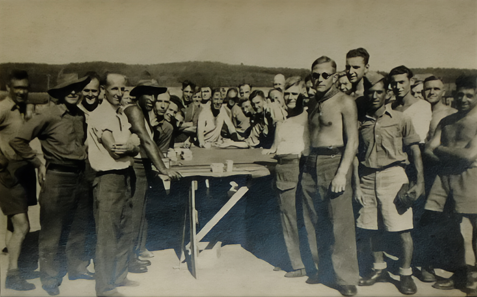 Large group of POWs standing closely together around a table outdoors, many smiling towards the camera.