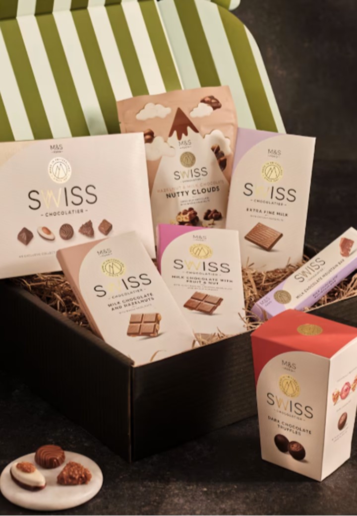 Gift box containing assorted Swiss chocolates, including bars, truffles, and nutty clouds, artistically arranged on straw with a striped lid. Luxurious and indulgent tone.