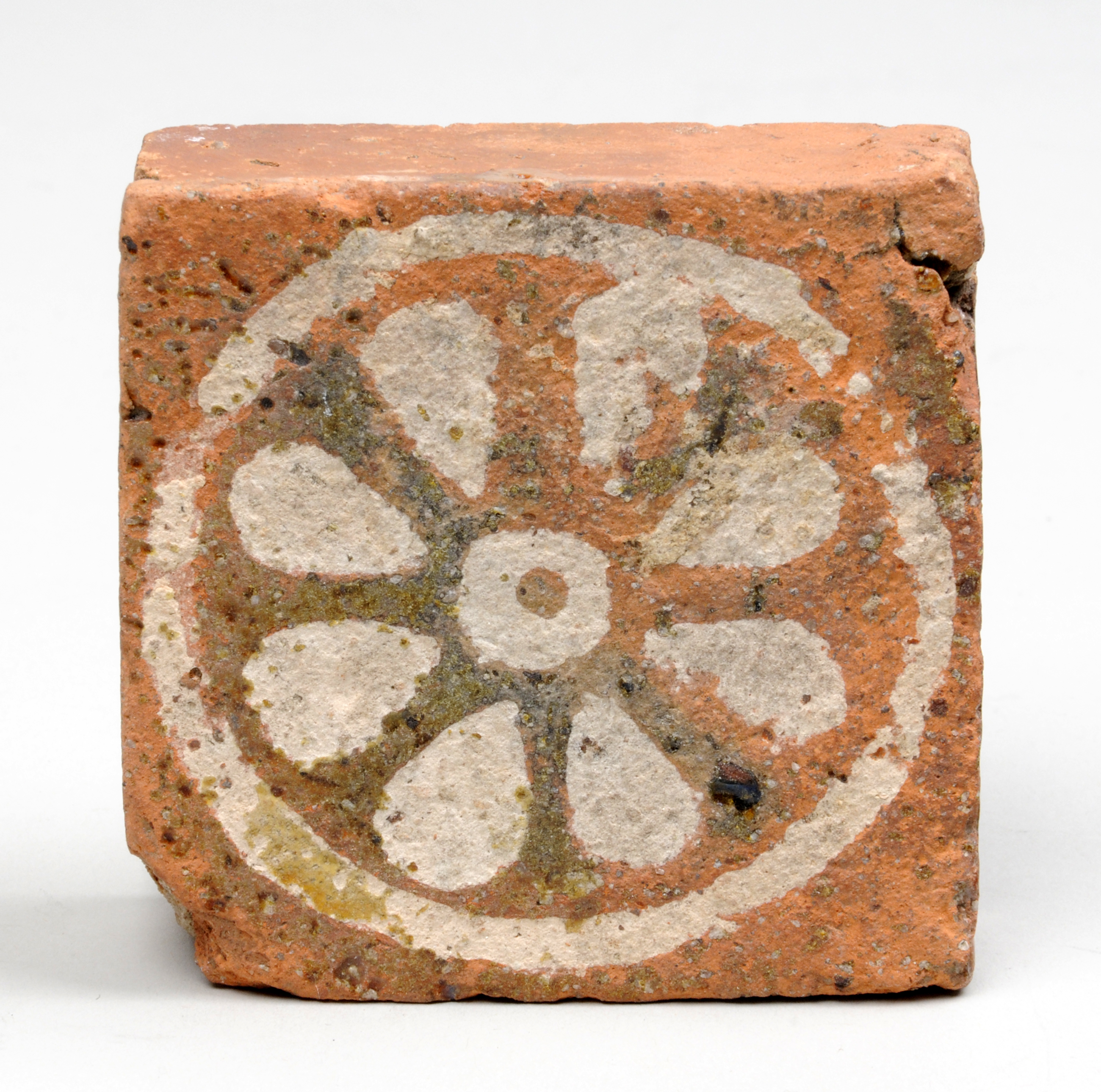 Colour photograph showing a square tile made of pinkish fired clay. It has a design on it in white of what looks like a flower in a circle