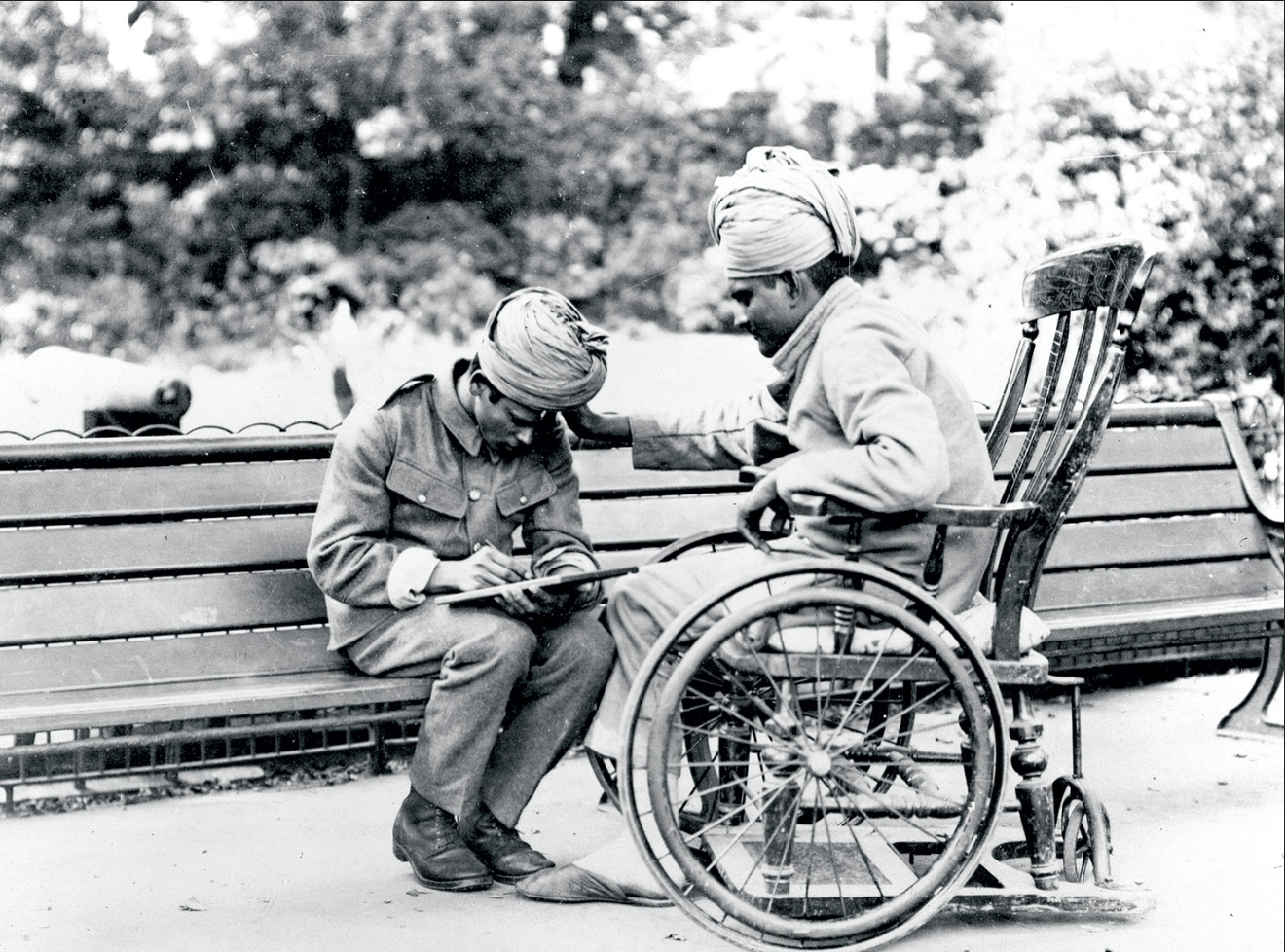 Black and white photograph of two Indian solders. One is in a wheelchair, the other is sitting on a bench. The one sitting on a bench is writing a letter.