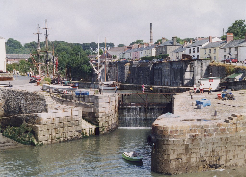 A colour photograph of a small harbour with what trickling down a small lock. People walk around. Boat masts are present to the left of the image and a row of houses are on the right.