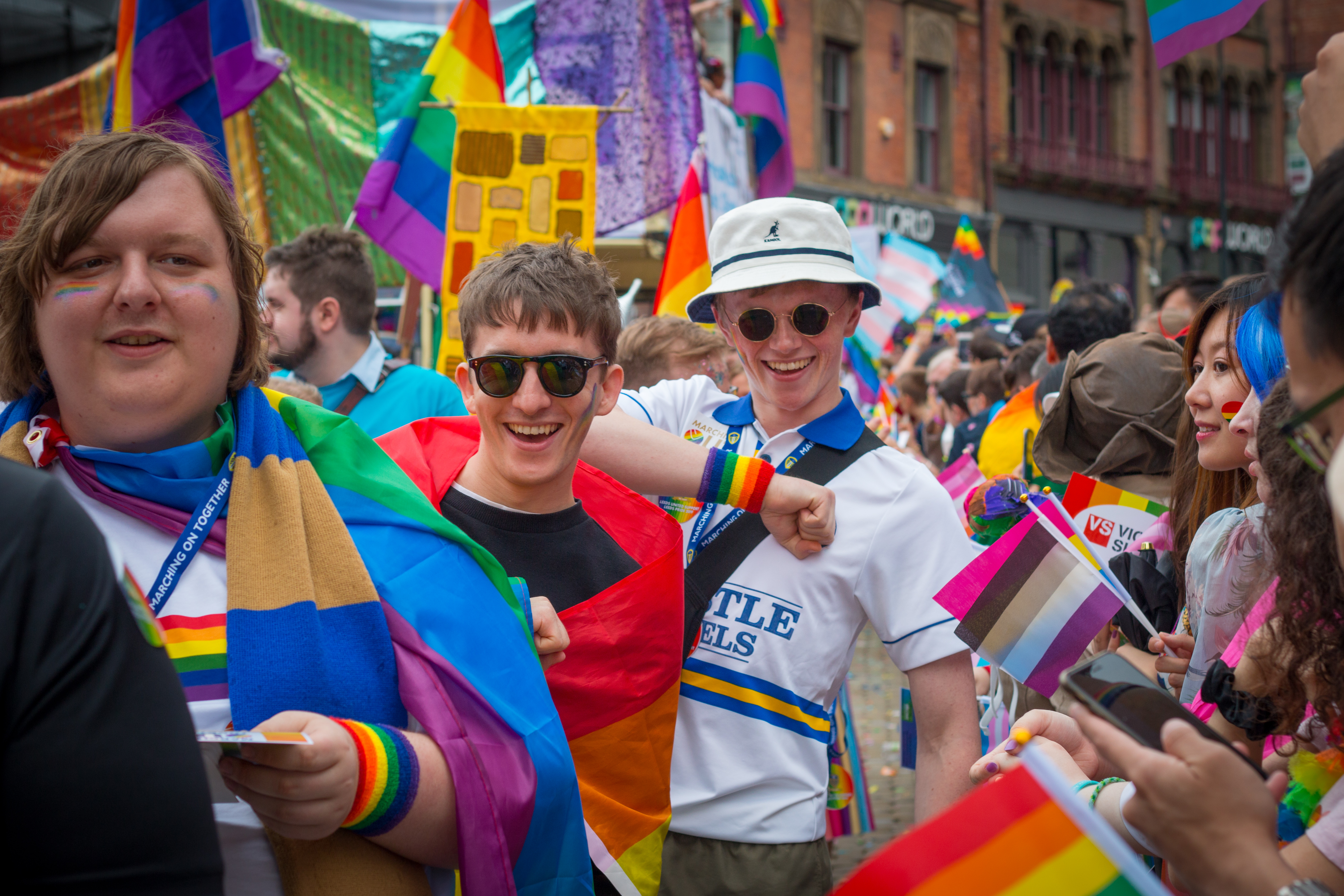 Colour photograph showing a close up of a crowd at Leeds Pride march wearing rainbow flags.  One is wearing a Leeds United shirt.