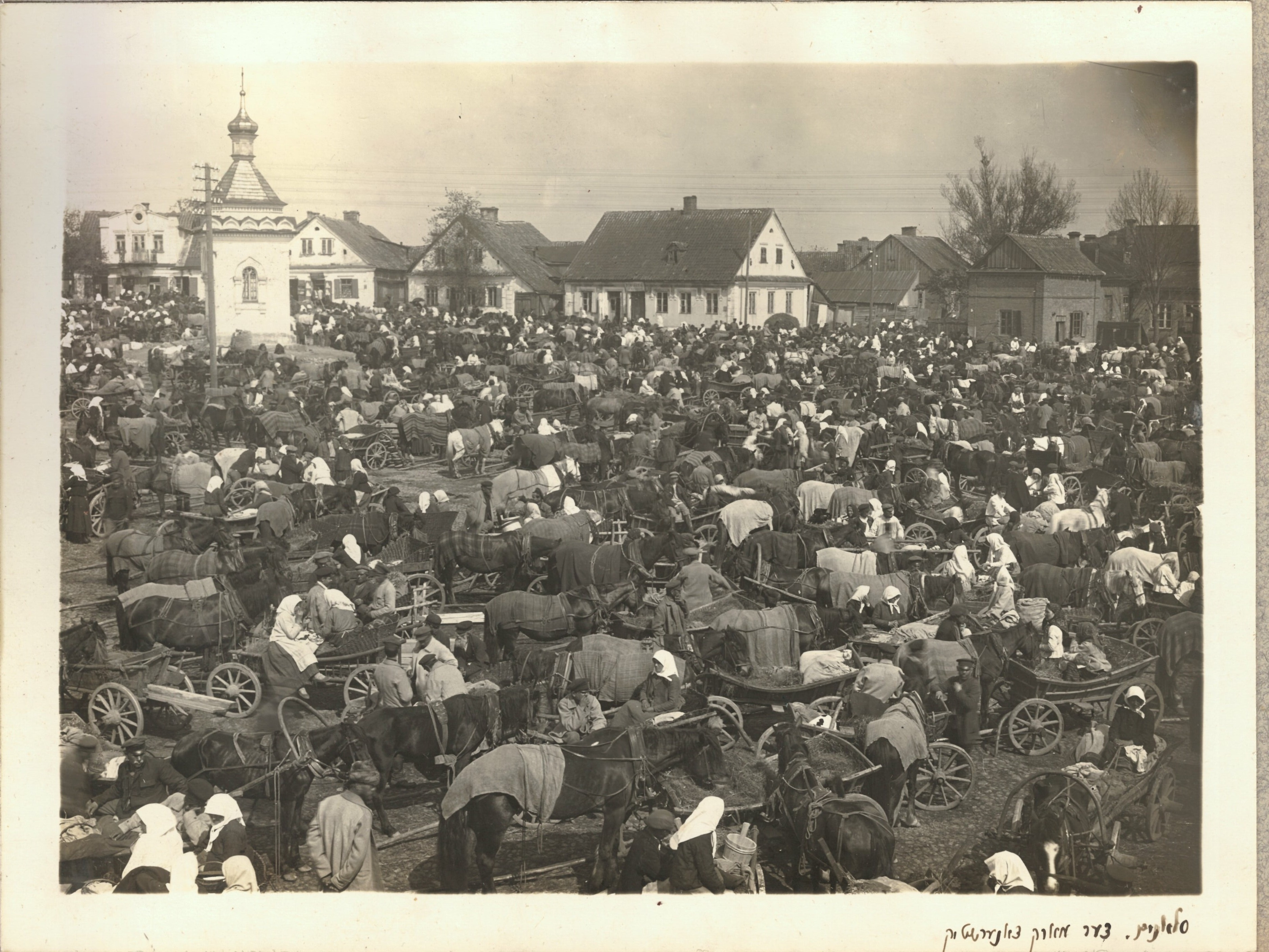 Black and white photo showing busy open air market with horses and carts bearing goods to sell and numerous people milling about