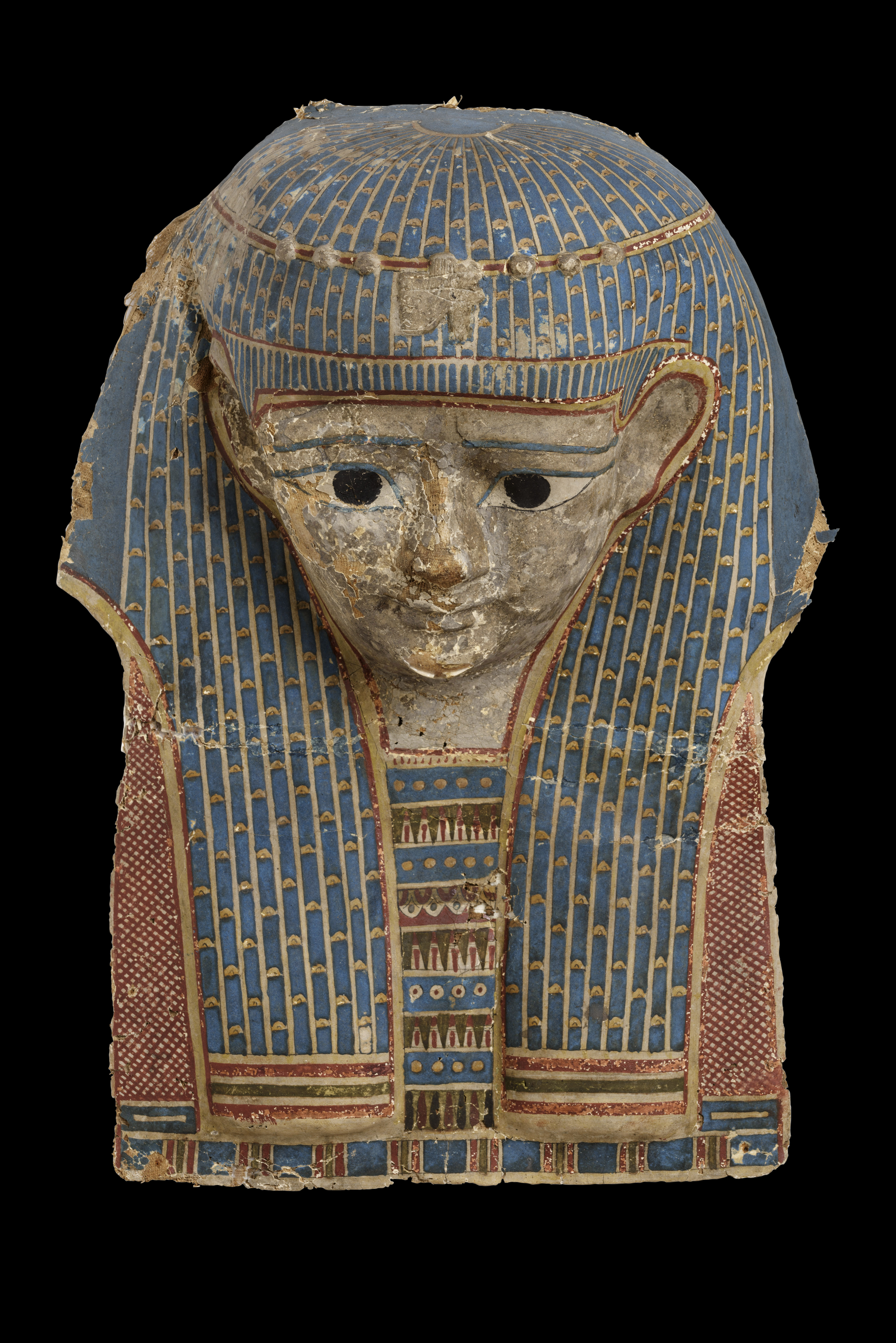 A painted ancient Egyptian cartonnage mask, featuring a face with large, black eyes and a calm expression. The mask is adorned with intricate designs, primarily in blue, red, and gold. The headdress has detailed vertical blue and gold stripes, while the shoulders are decorated with geometric patterns. The mask shows signs of age, with visible cracks and wear on the surface.