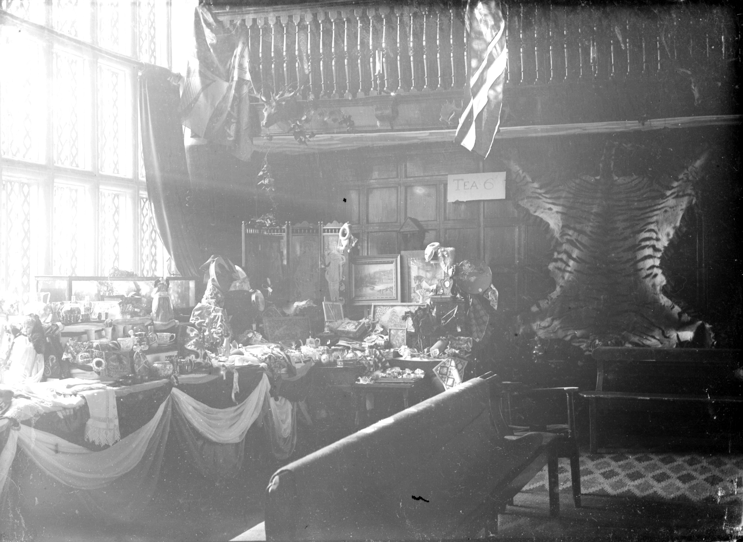 Black and white photo of a room with a lit display table showing assorted items, possibly for sale. A "Tea 6" sign is visible, alongside a mounted tiger skin.