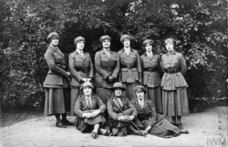 A black and white photograph of nine women in uniform pose outdoors against dense foliage, some standing, others seated. They wear military-style attire and caps.