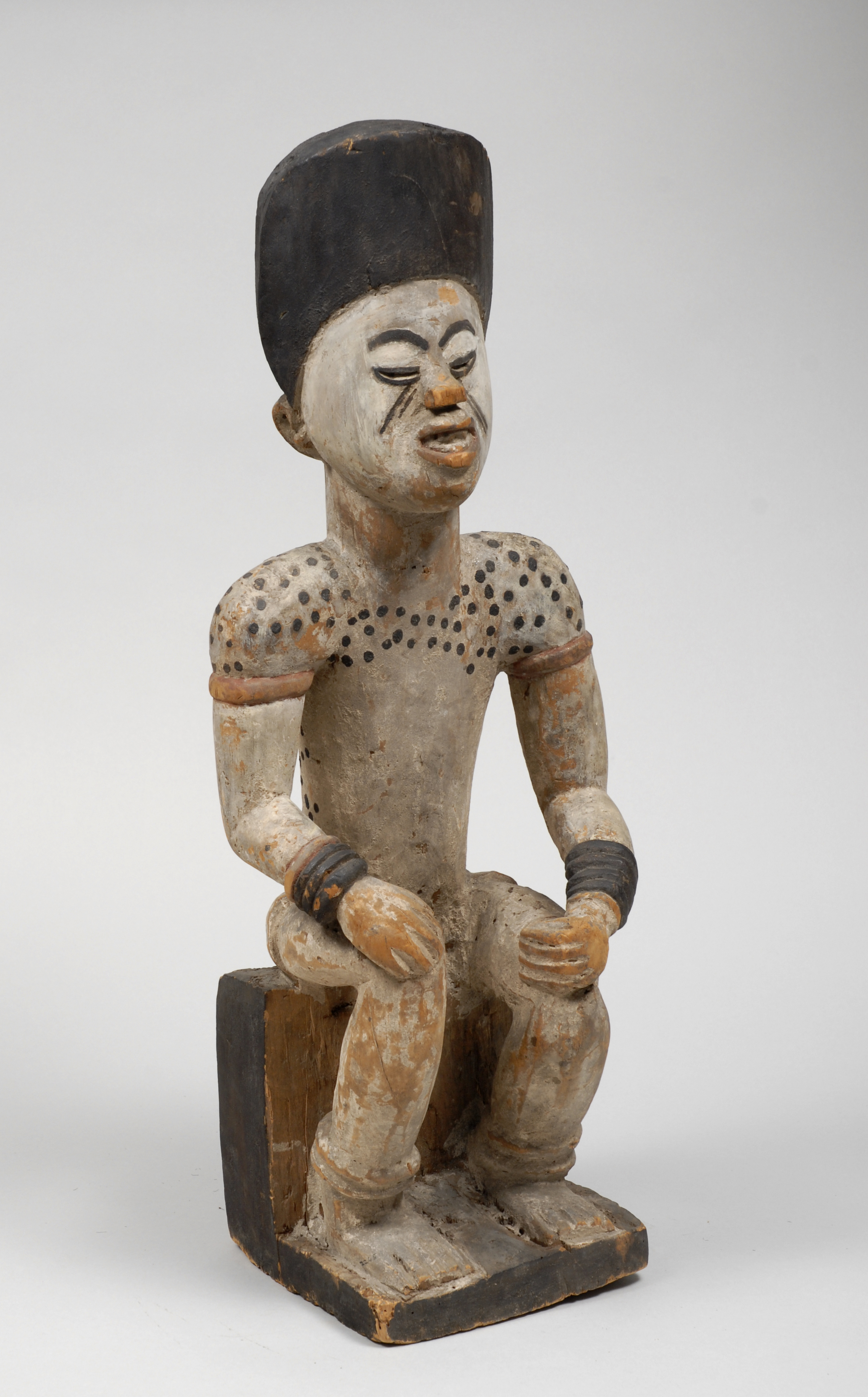 A wooden sculpted figure of a man sitting down with his hands on his knees. His shoulders have a black spot pattern.  He has black bangles on both wrists, and tall black painted hair. The rest of the sculpture is brown wood colour.