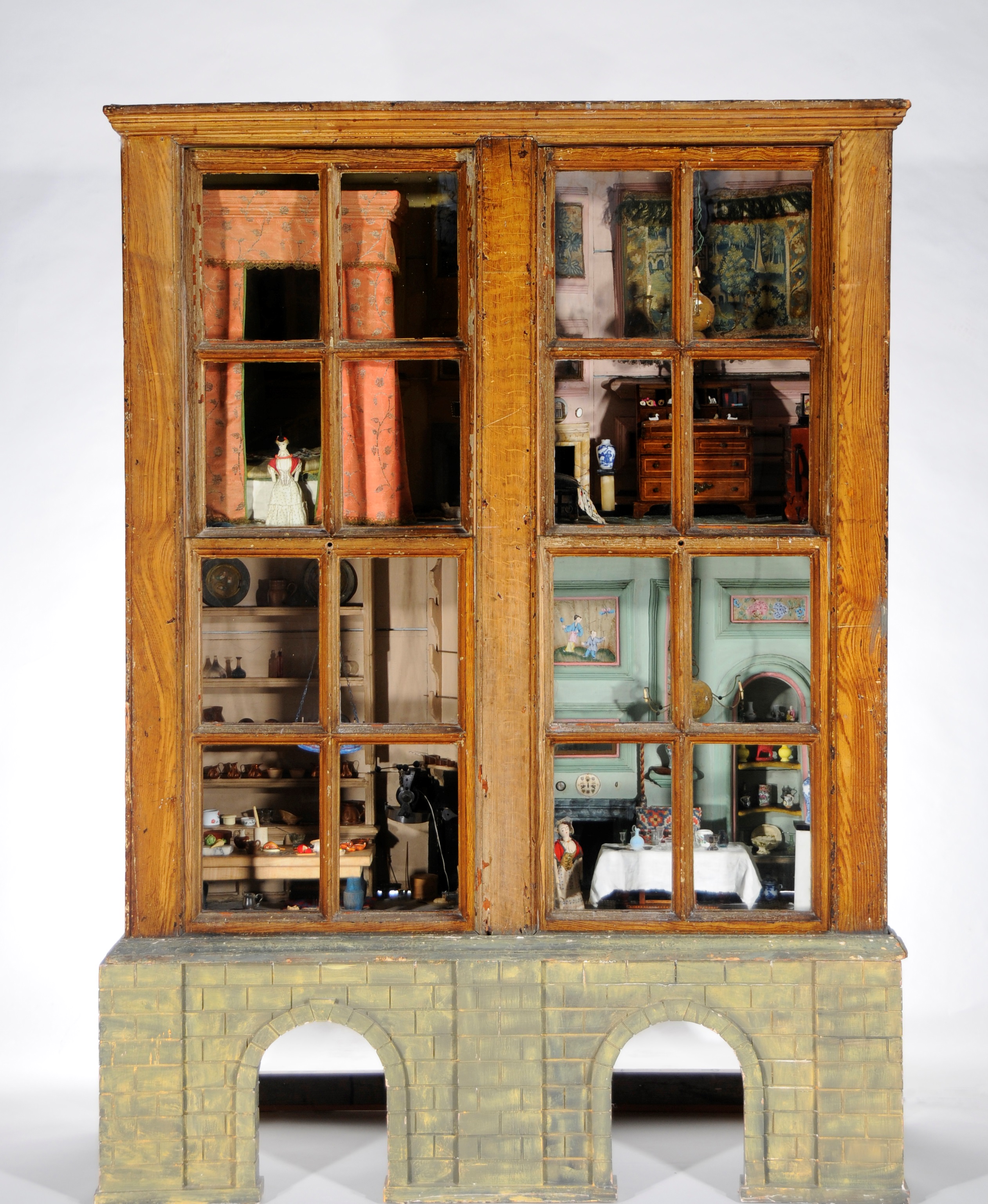 A wooden cabinet style 'baby house' from the 1750s, on a more recently added base that has been painted with a brick pattern. The interior has two floors, with two highly decorated and furnished rooms on each floor. These include a bedroom with curtained four-poster bed, a kitchen with crockery on shelves and food on the table, and light fittings from the ceiling.