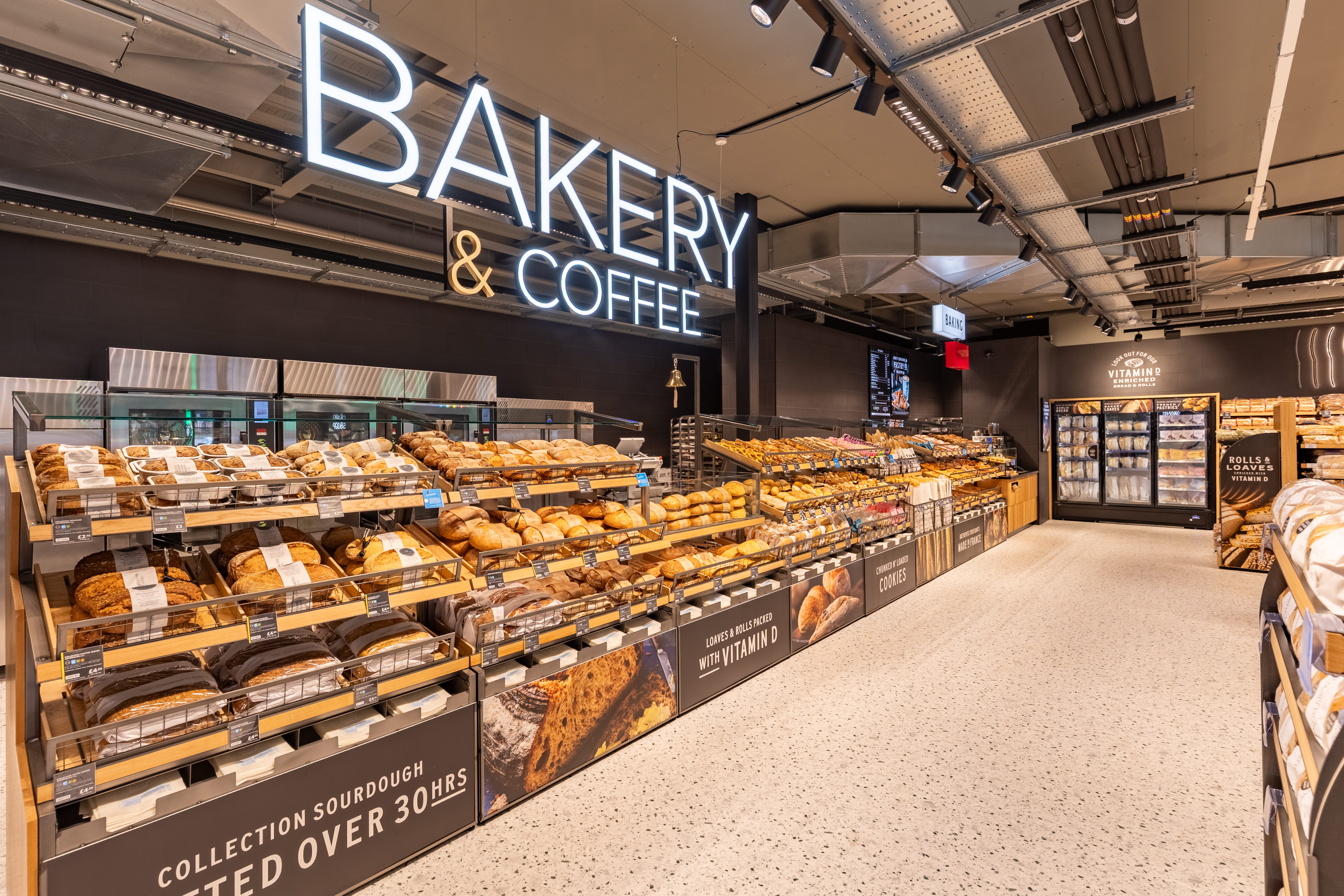 Modern bakery section in a Marks & Spencer supermarket, with various bread and pastries on wooden shelves. A “Bakery & Coffee” sign is visible above, creating a warm, inviting atmosphere.
