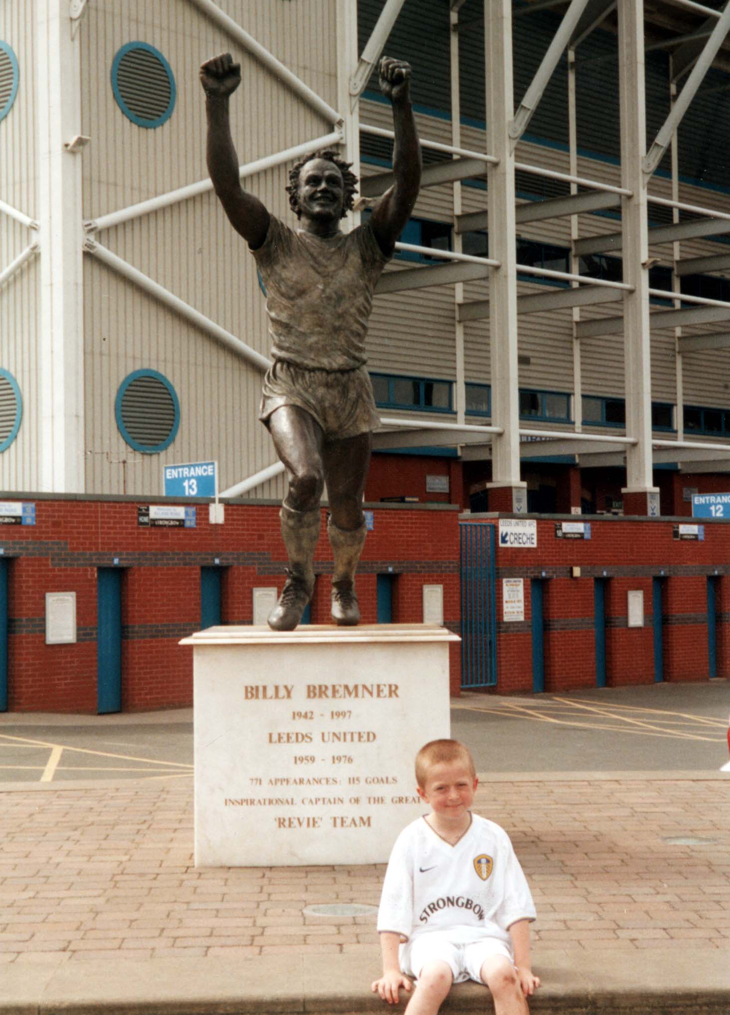 Photo of statue of Billy Bremner outside Leeds United football ground. The statue has his arms raised and is made of bronze. A small boy in a white Leeds United kit sits in front of the statue.