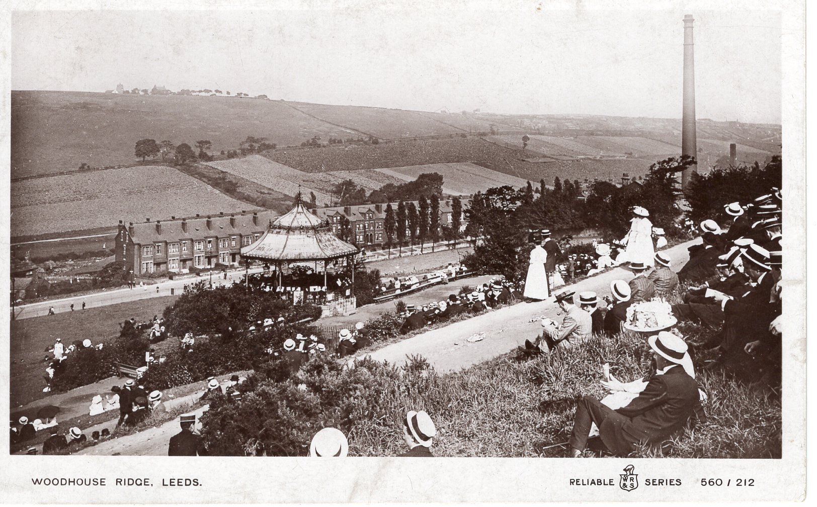Black and white photograph showing a view down a hill towards a road at the bottom with houses. There are fields on the other side of the road. Down the hill, there are people sitting on the grass between paths running horizontally across the image