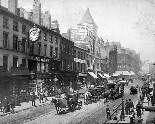 Black and white photograph showing the Briggate area of Leeds.  There are lots of people in the wide street, and horses and carriages