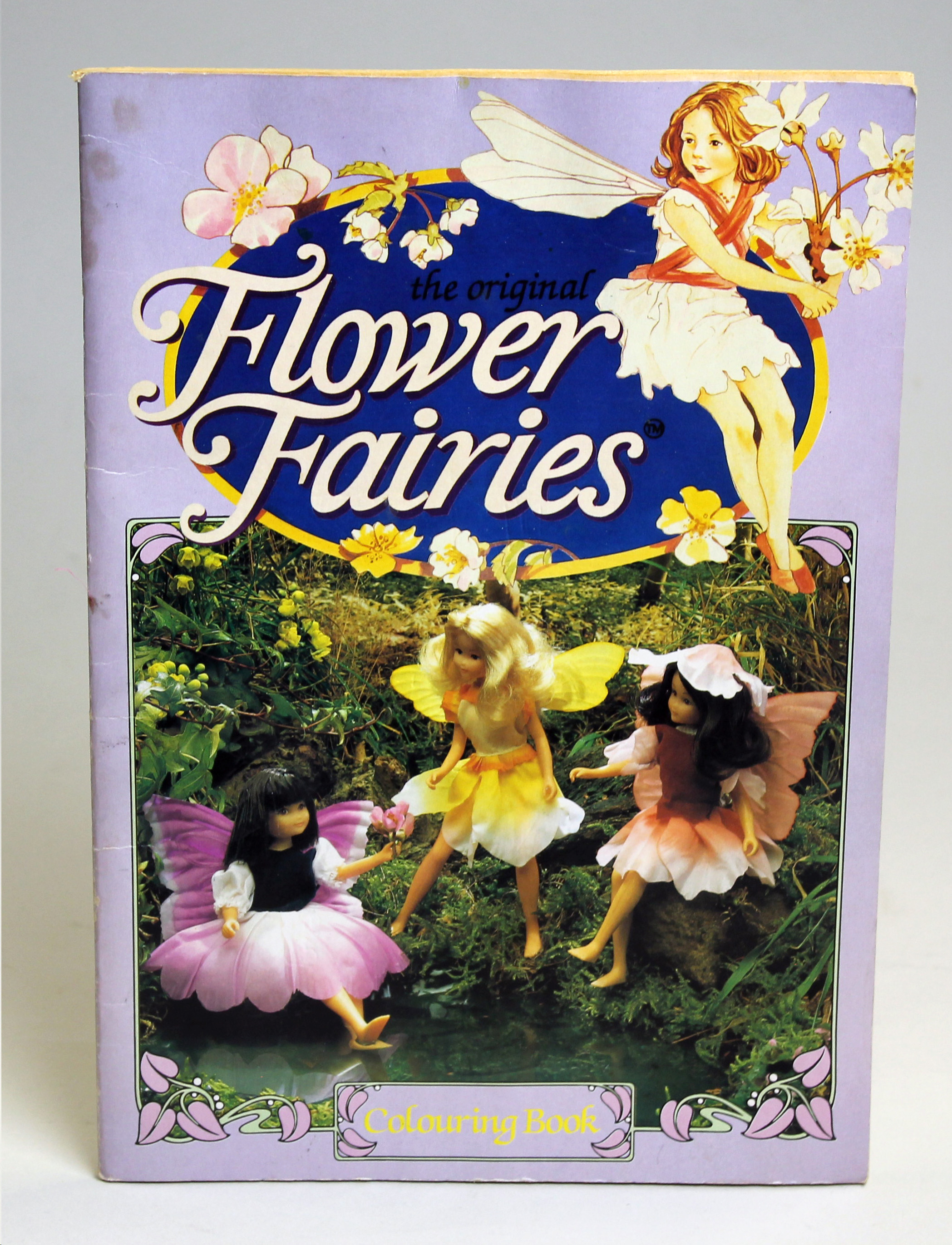 Colour image of the front of a Flower Fairies colouring book. The cover image has a photo of three flower fairy dolls sitting by a pond