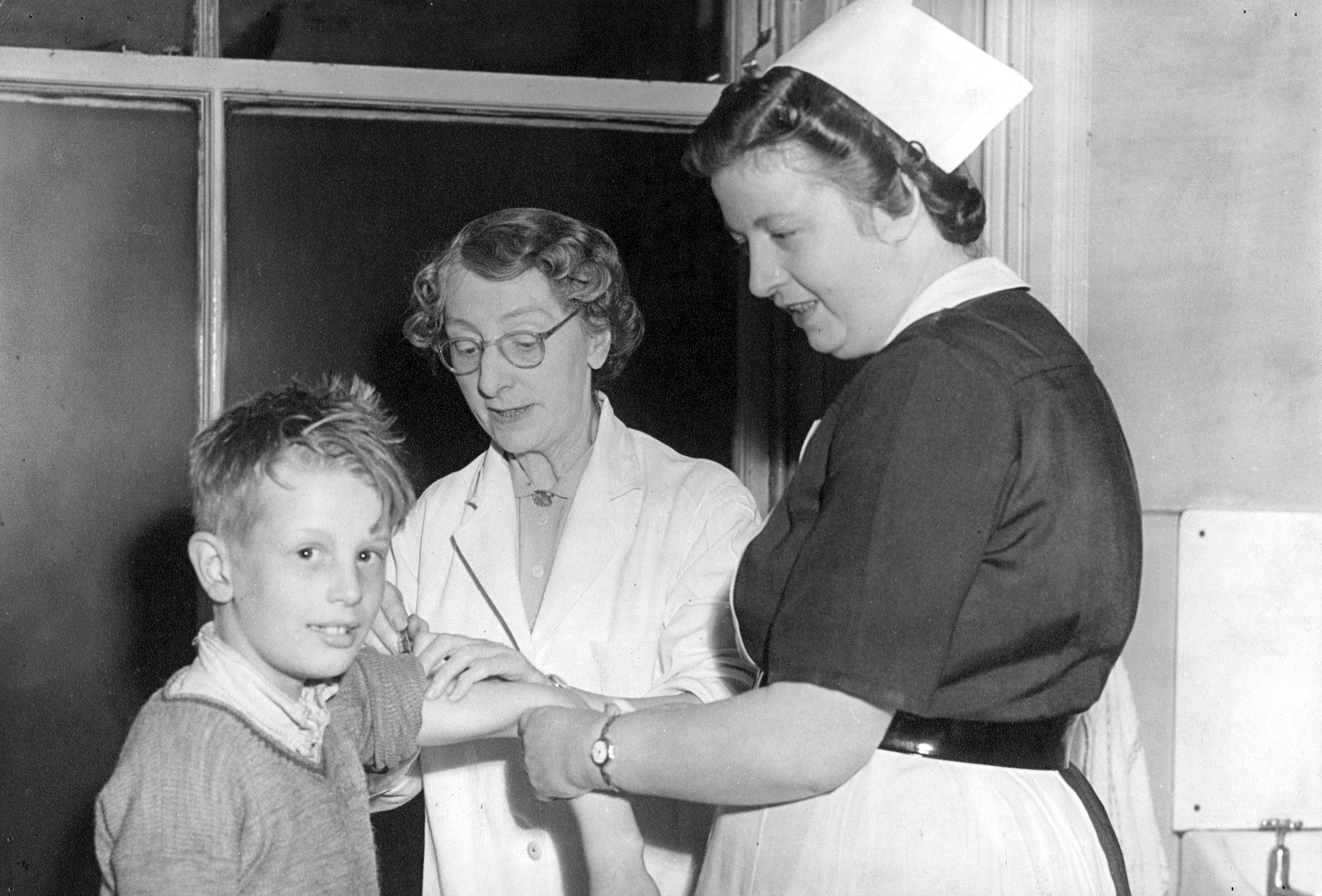 A female doctor in a white coat with glasses, administers a vaccine to a young boy, who has an apprehensive smile towards the camera. A nurse smiles while supporting the boy's arm. The setting is a medical office.